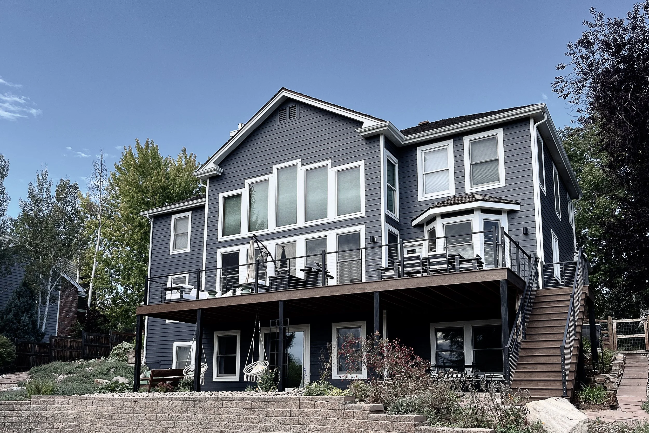 A multi-story house painted in gray with white trim, featuring numerous windows and a large balcony with outdoor furniture. The house is set against a blue sky with some trees nearby and a stone garden border in the foreground.