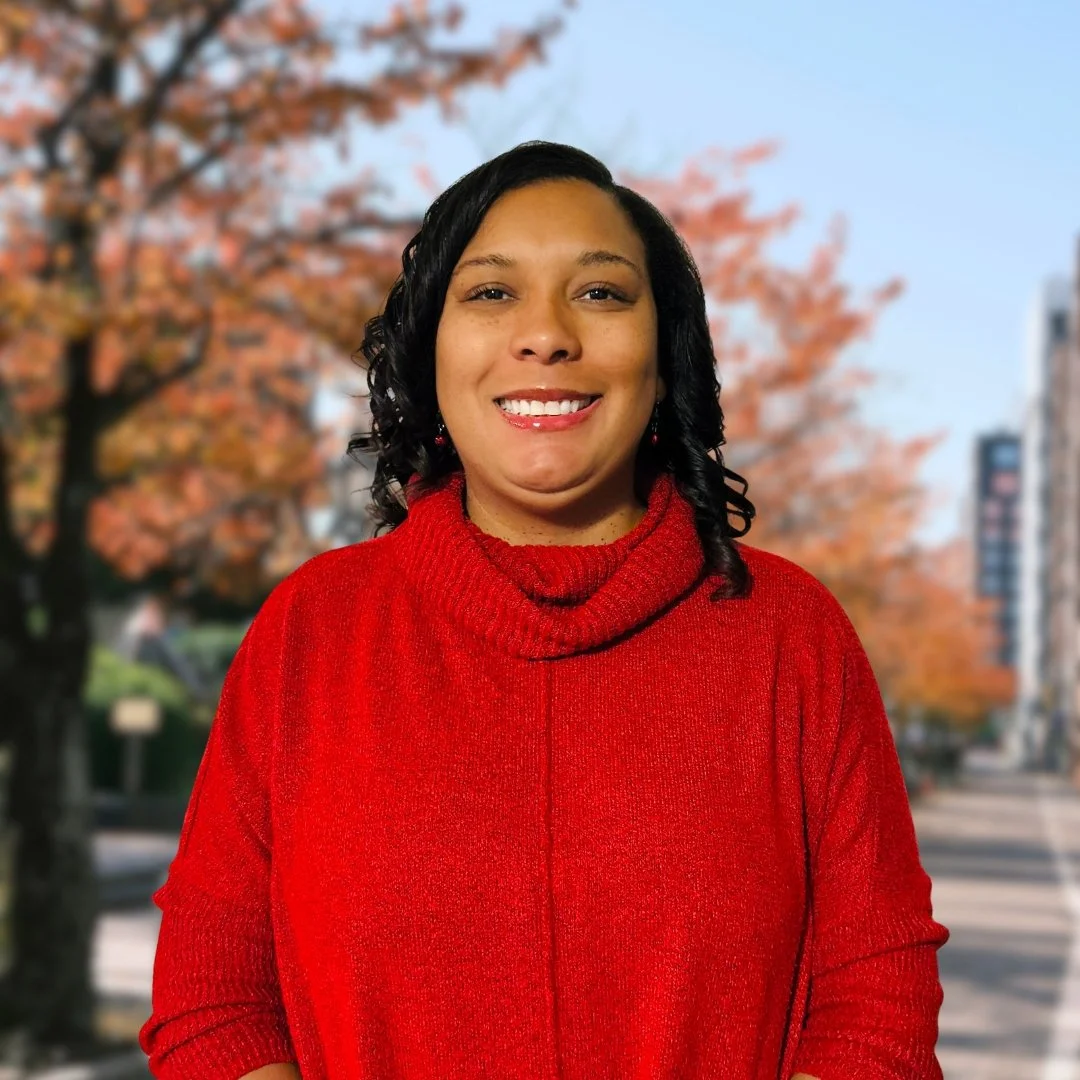 Woman smiling outdoors in front of trees with fall foliage, wearing a red turtleneck sweater.