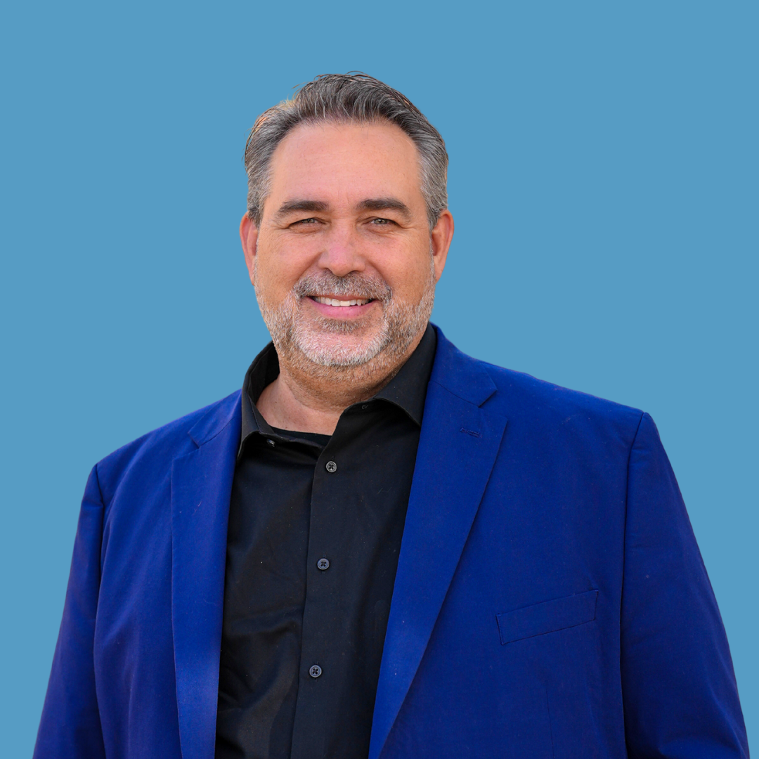 A middle-aged man with gray hair and a beard, dressed in a black shirt and a blue blazer, smiling against a light blue background.