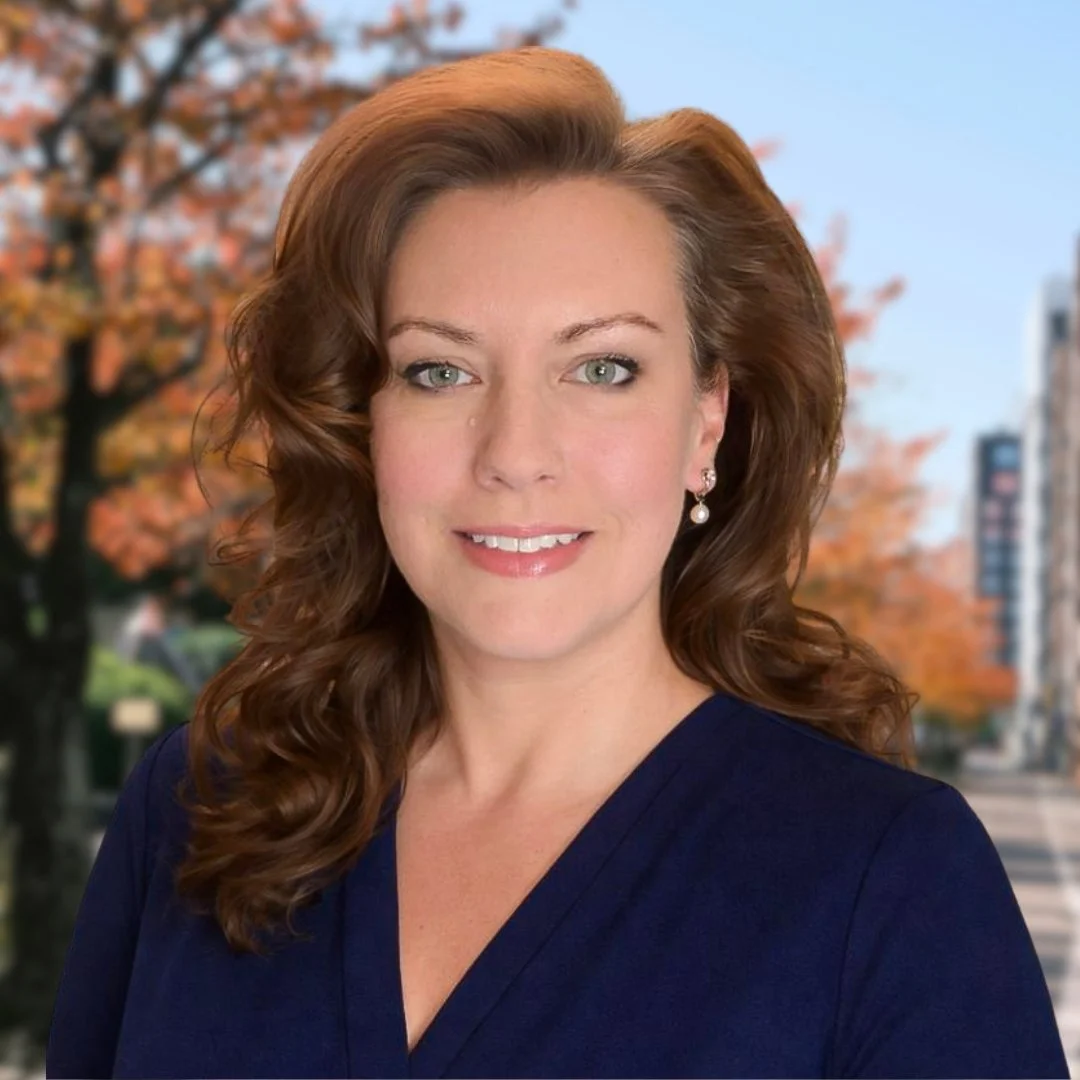 A woman with long, curly auburn hair, light skin, and green eyes, wearing pearl earrings and a navy blue top, standing outdoors with autumn trees and city buildings in the background.