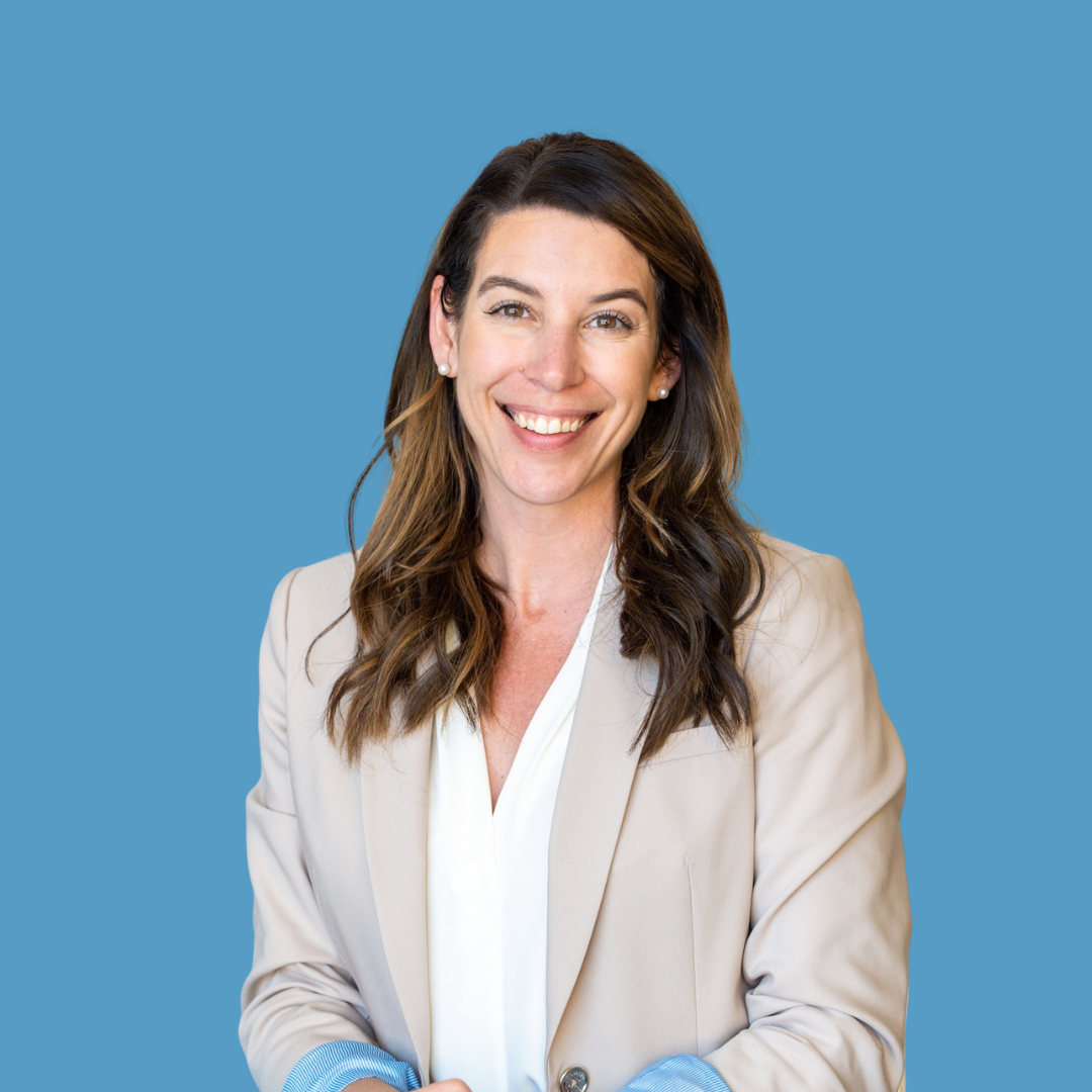 Portrait of a smiling woman with brown hair, wearing a beige blazer and white shirt, against a blue background.