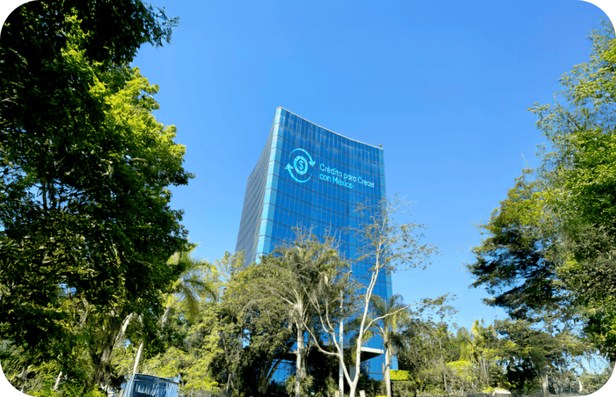 Rascacielos de cristal con un mensaje en español sobre crédito para crecer con México, rodeado de árboles y cielo azul.