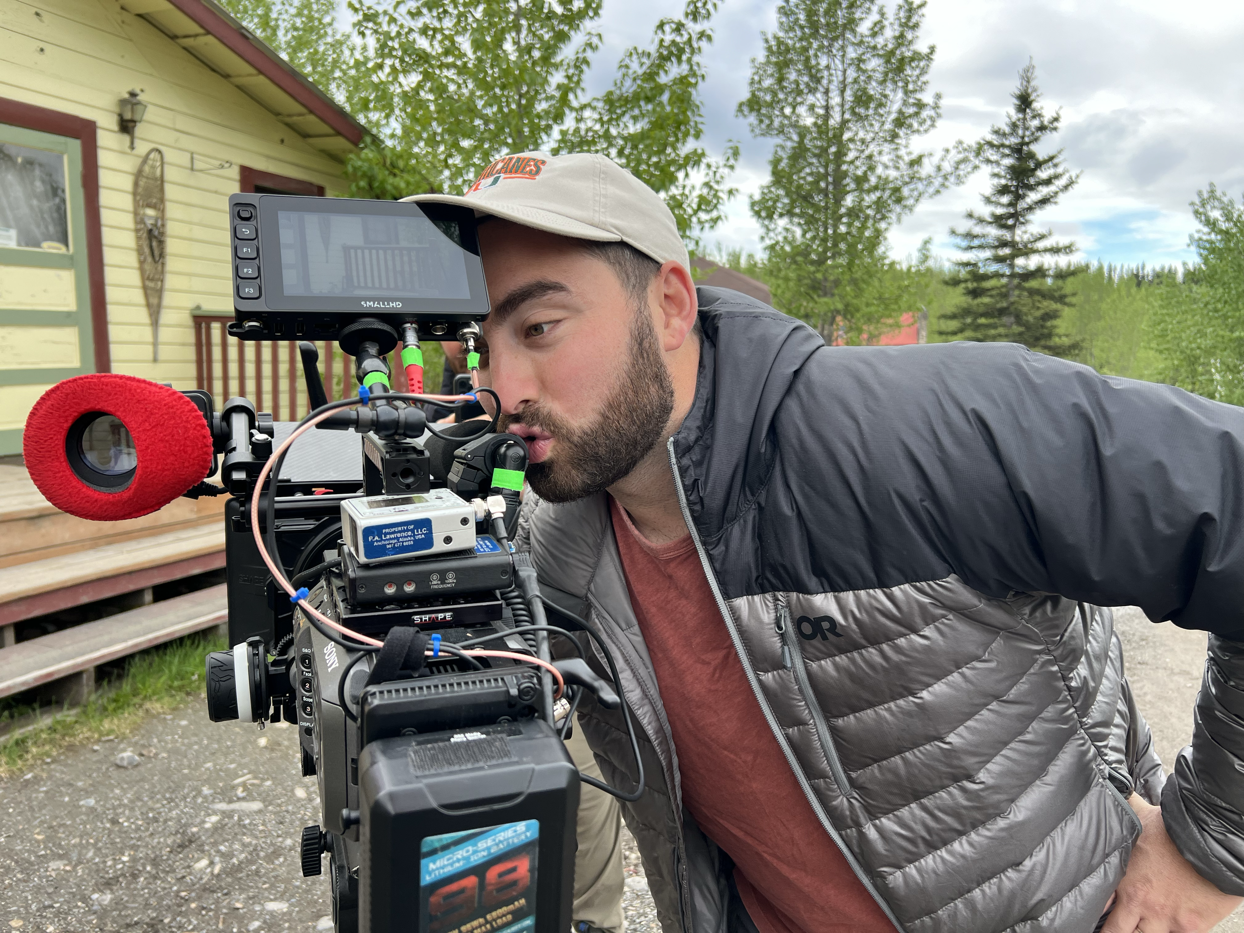 A man operating a professional film camera outdoors, with a house, trees, and cloudy sky in the background.