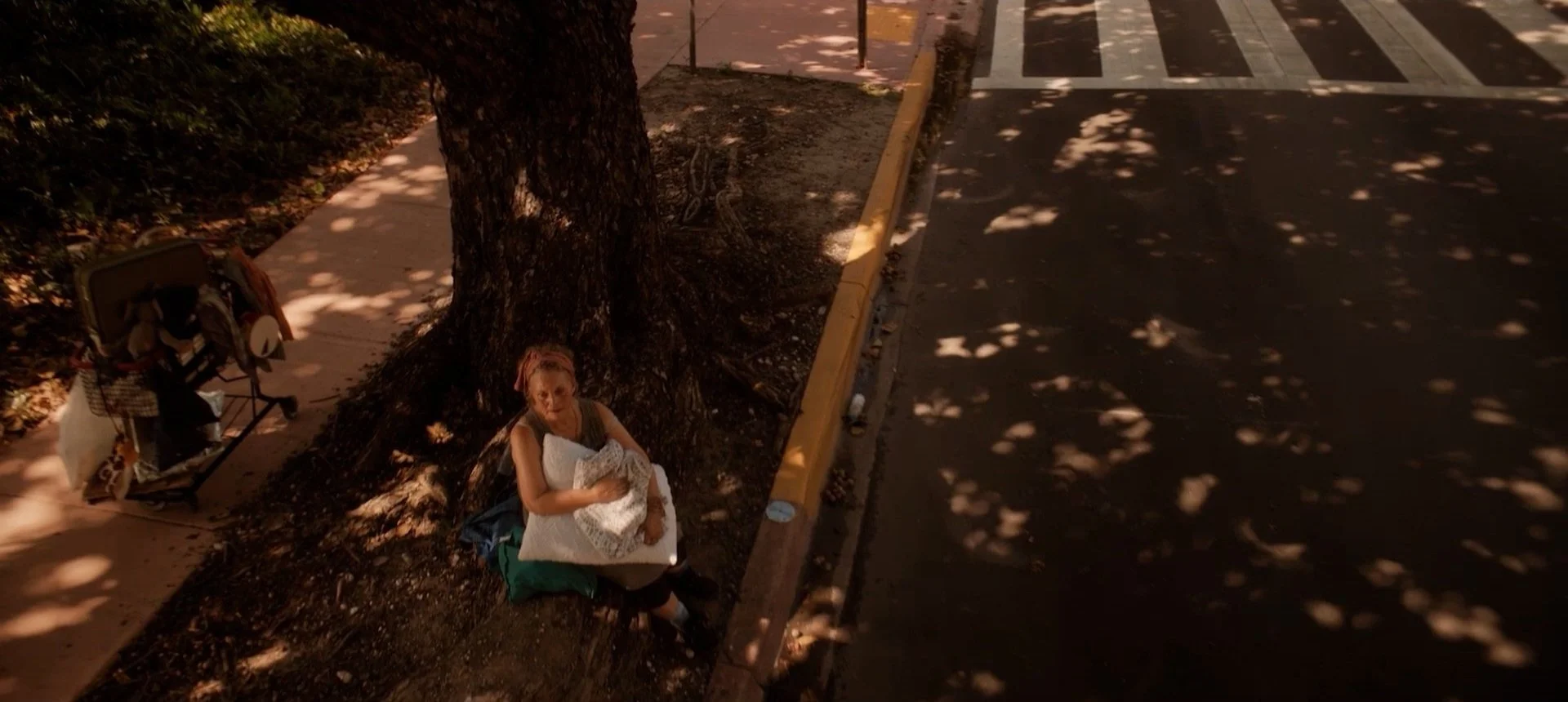 A person sitting under a tree on a sidewalk, holding a baby wrapped in a blanket. Nearby, a cart filled with belongings is parked. Sunlight creates dappled shadows on the ground and street.