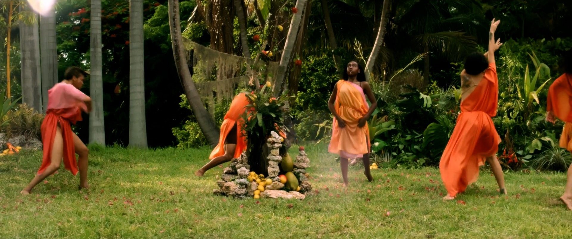 People in colorful dresses dancing around a stone altar with fruits, in a tropical garden.