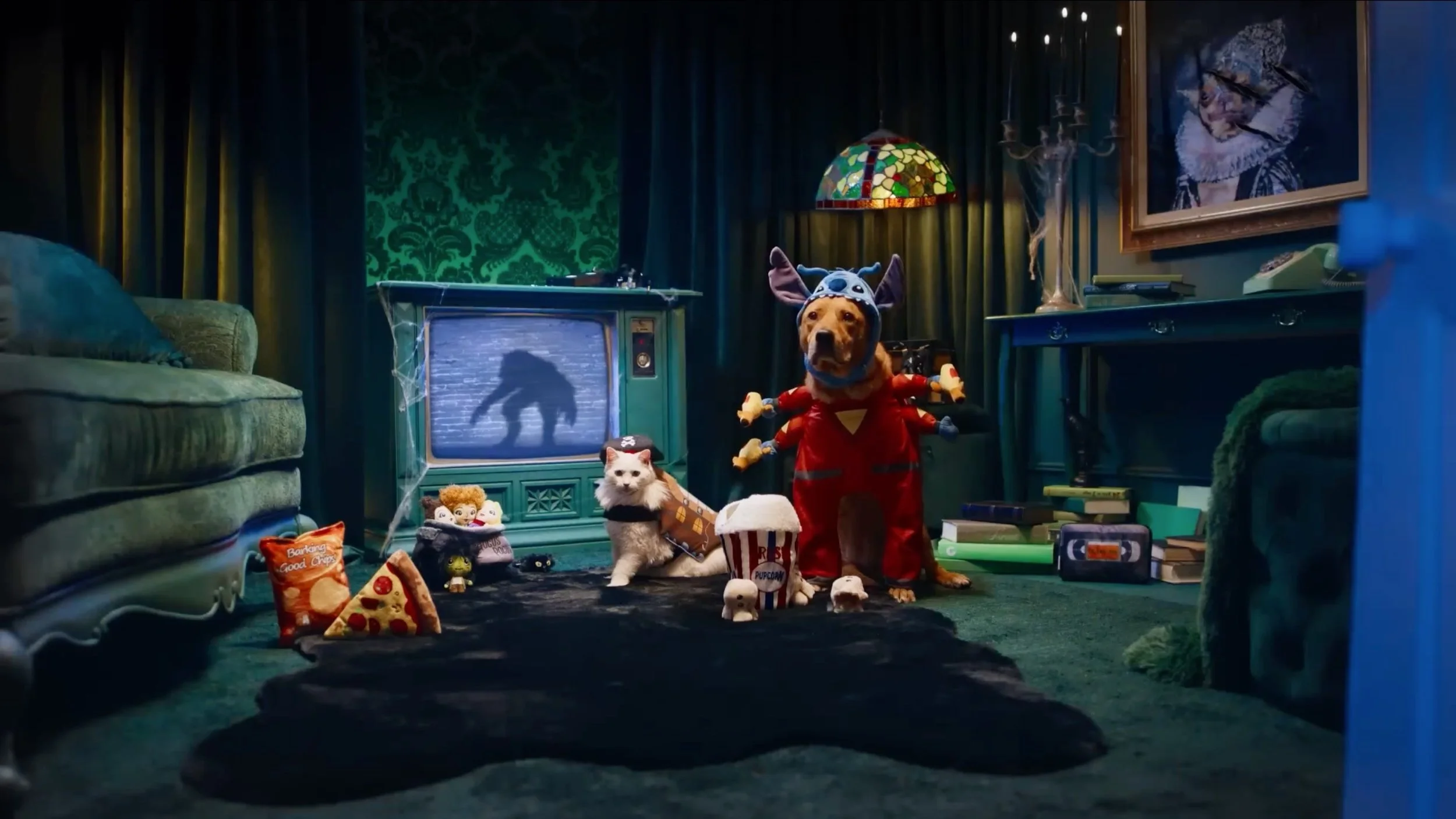 A dog dressed as a superhero with multiple arms, standing in a cluttered living room with a second dog sitting nearby, surrounded by popcorn, snacks, and toys, with a TV showing a shadow of a creature.