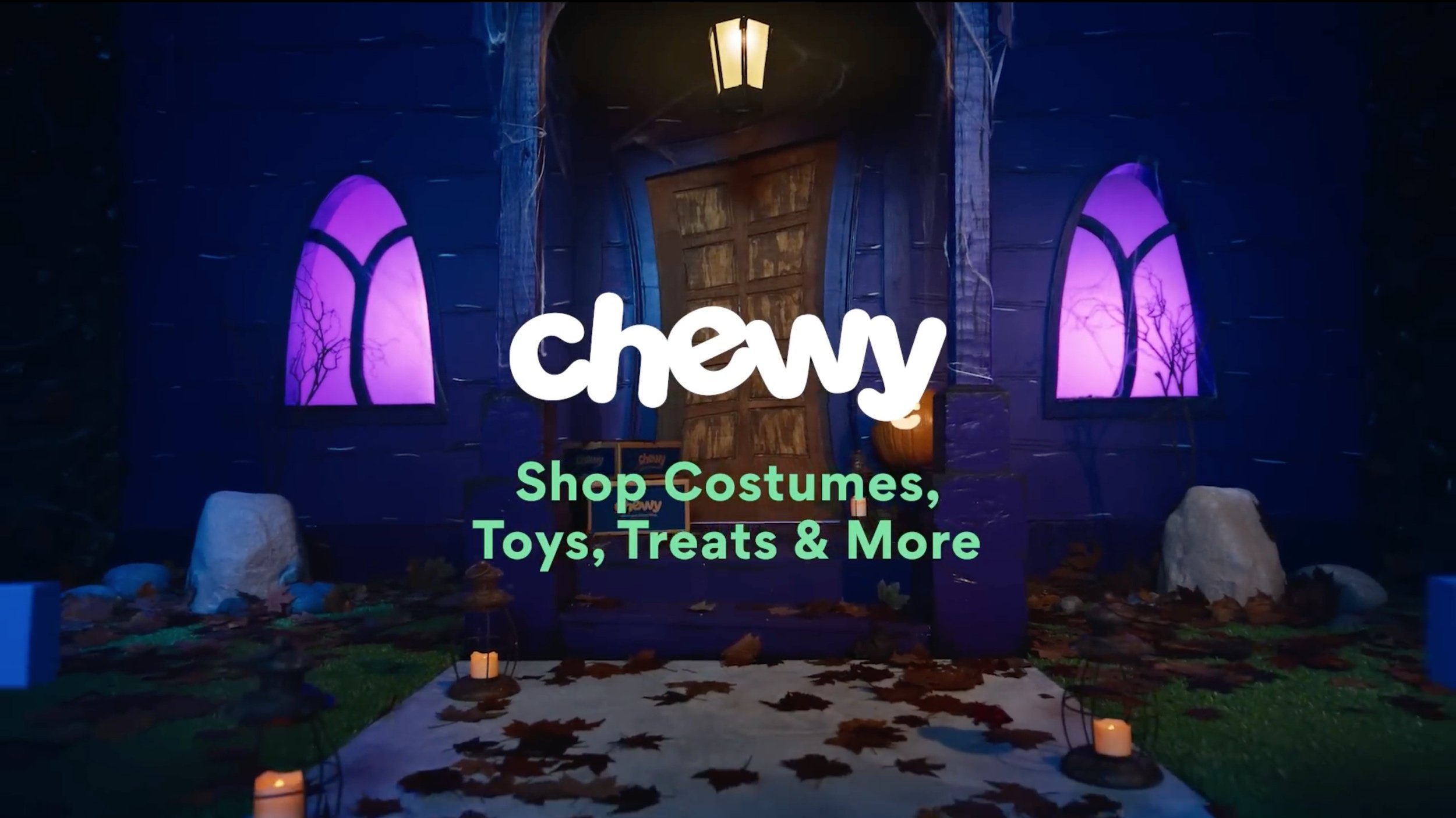 A Halloween-themed miniature house with purple windows, a wooden door, and orange candles illuminating a pathway covered with fallen leaves. The scene is decorated with faux rocks and spooky trees in the background.