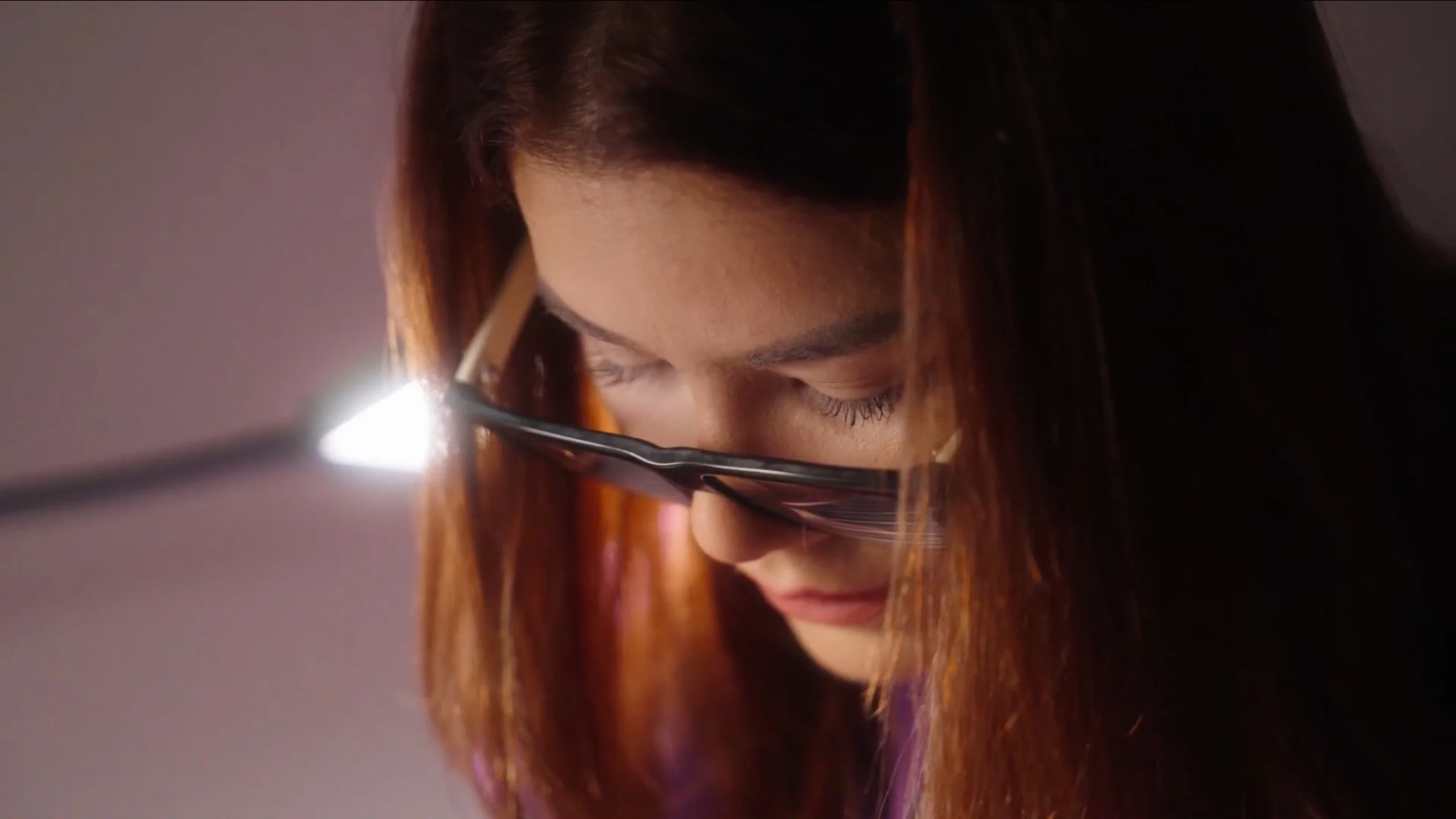 A woman with long auburn hair wearing glasses and looking down in a dimly lit setting with a bright light illuminating her face.
