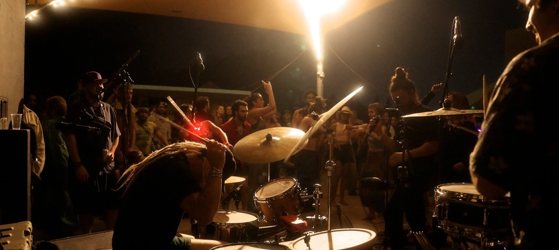 A live music performance with a drummer in the foreground, a singer with dreadlocks leaning over the drums, and a crowd of people dancing and enjoying the night scene in a dark setting illuminated by warm overhead lighting.
