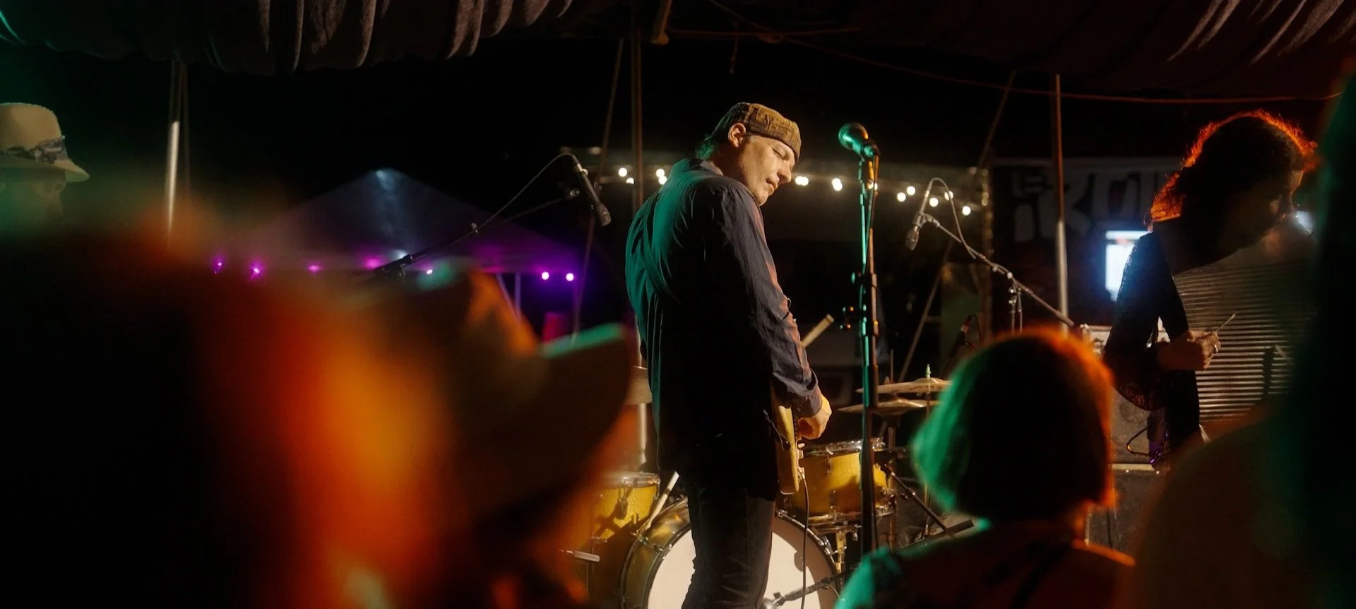 Musicians performing on stage at a dimly lit venue, with one person playing the guitar and a woman looking at her phone.