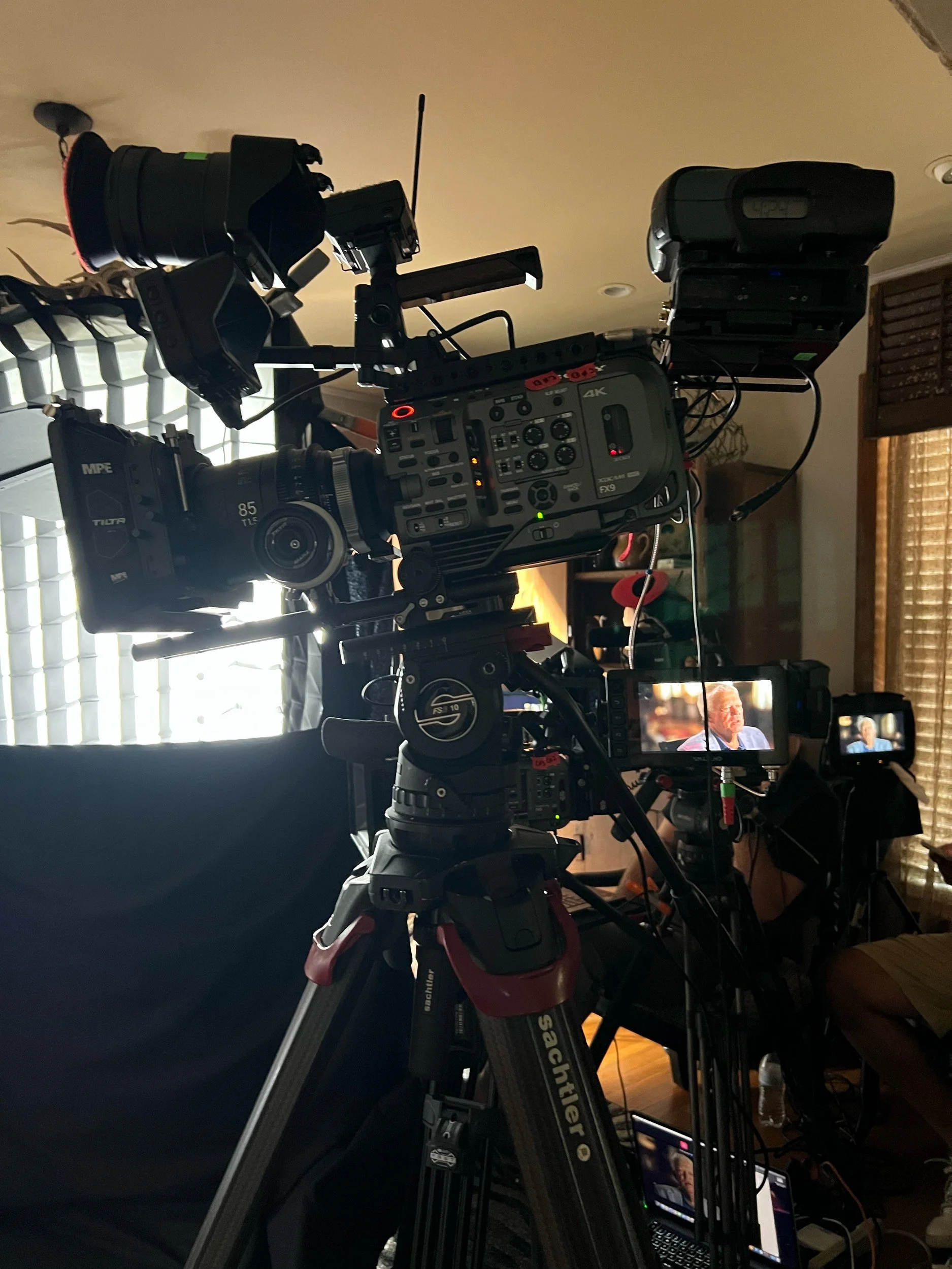 Professional video camera on tripod filming an interview indoors, with two monitors displaying a woman speaking.
