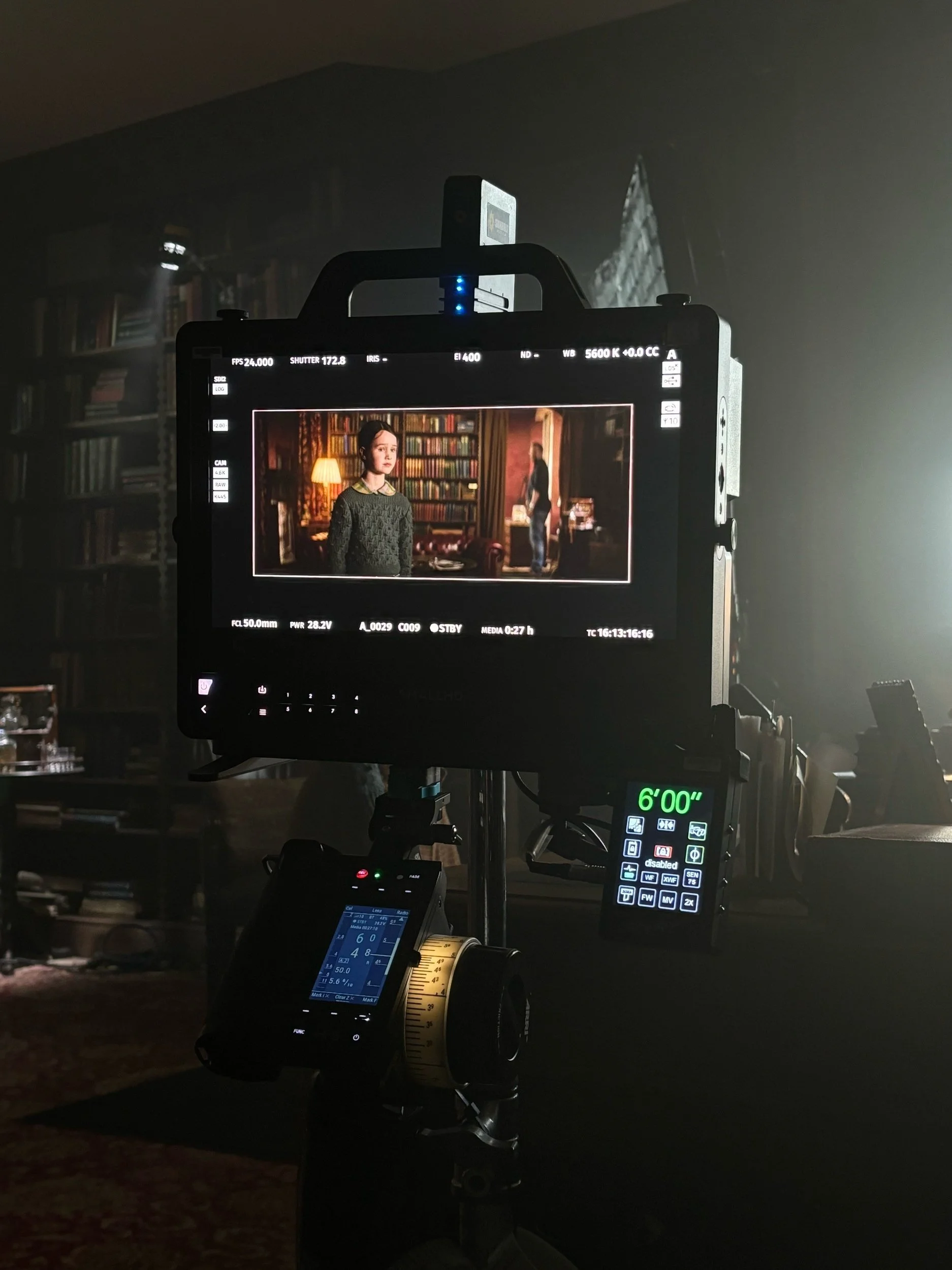 A film set with a monitor showing a scene from a movie or TV show featuring a woman with a bookshelf in the background, supported by filming equipment including a follow focus and remote control.