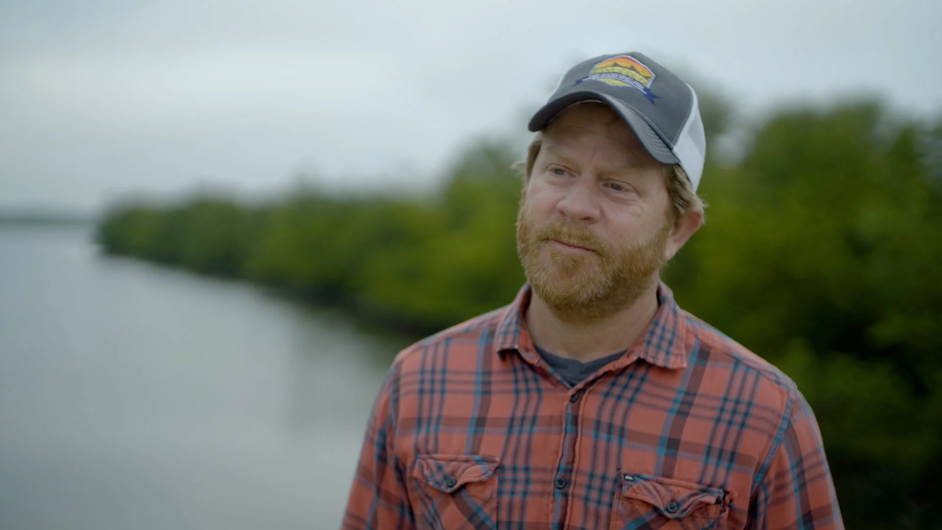 Man in a plaid shirt and cap standing outdoors near a body of water with trees in the background.