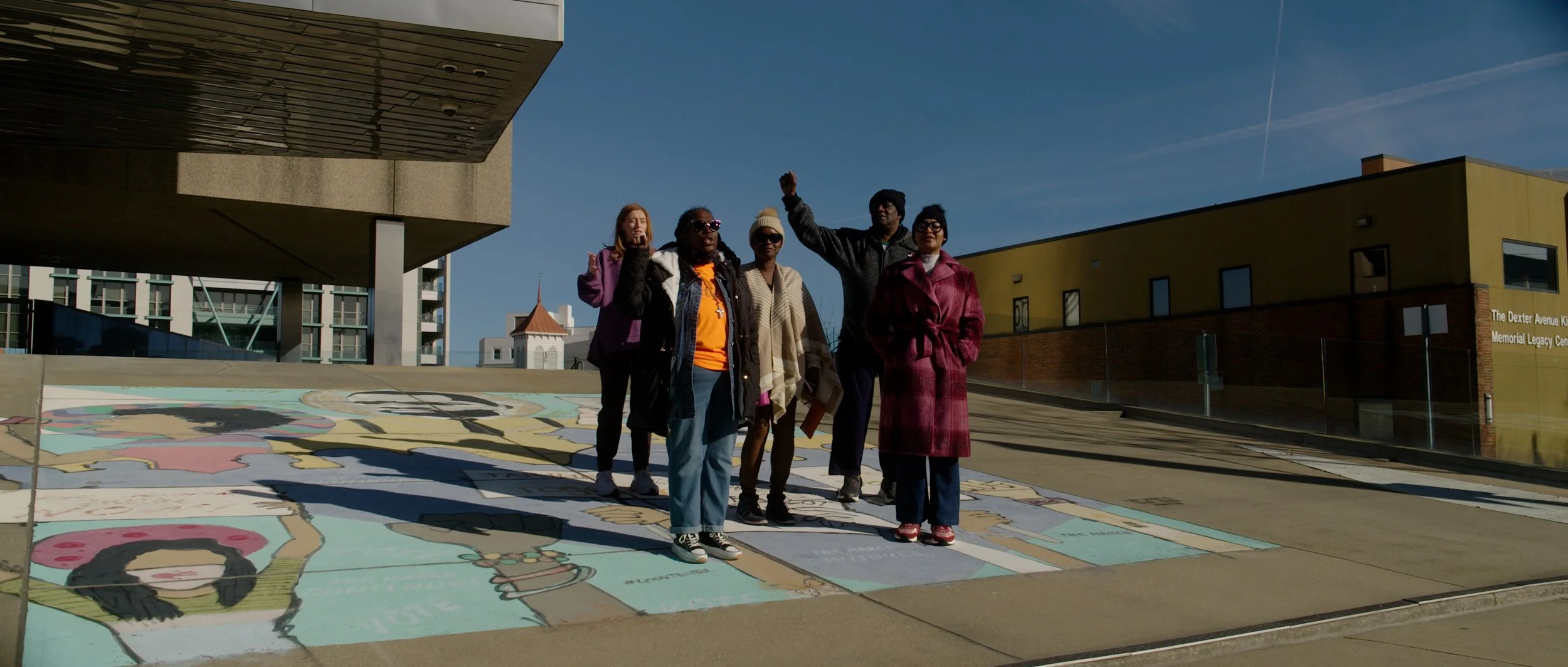 Group of six people standing on a colorful mural painted on pavement outdoors, with buildings in background and clear blue sky.