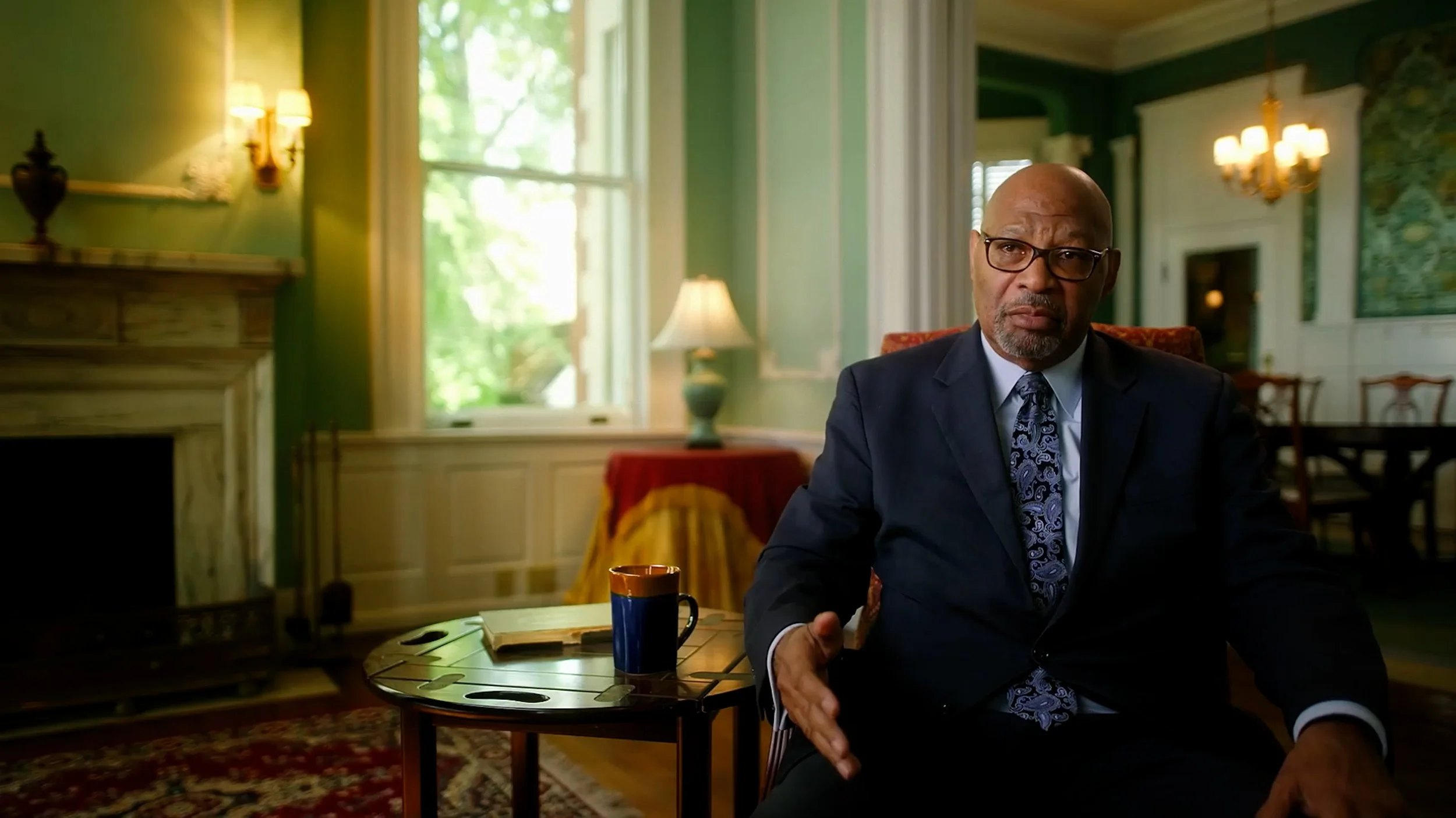A man in a suit sits in an elegant living room with green walls, a chandelier, and a large window. A fireplace and table with a mug and book are in the foreground.