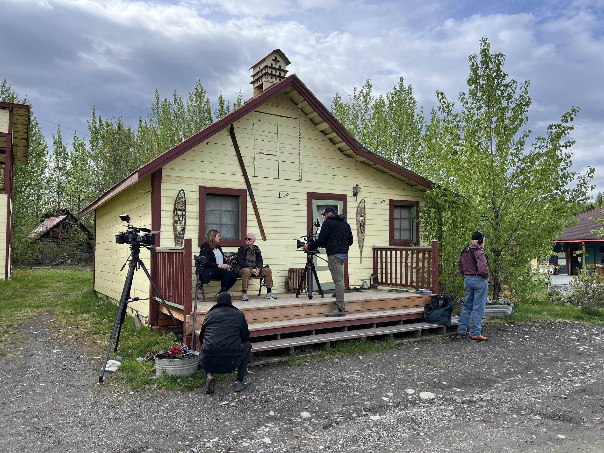 Filming crew conducting an interview outside a yellow house with red trim, with camera equipment set up on a wooden porch, and trees and buildings in the background.