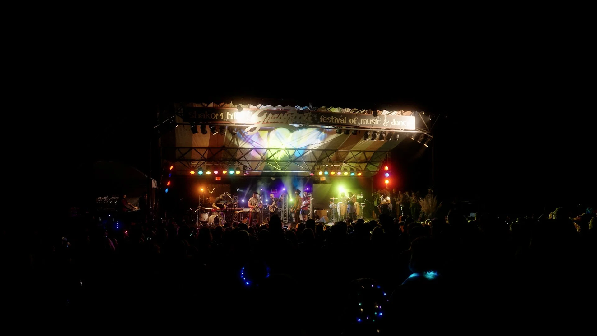 Live music performance on an outdoor stage at night, illuminated with colorful stage lights, with an audience in front of the stage.