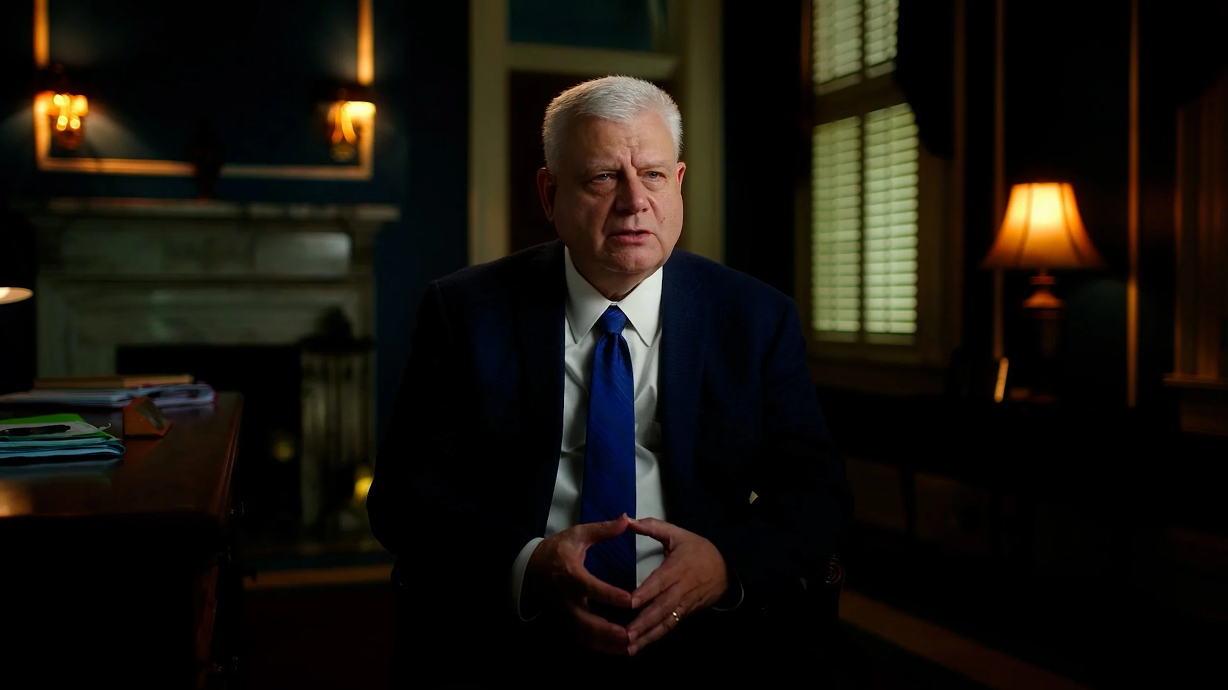 A man in a suit and tie sitting in a dimly lit room with lamps and a fireplace in the background.