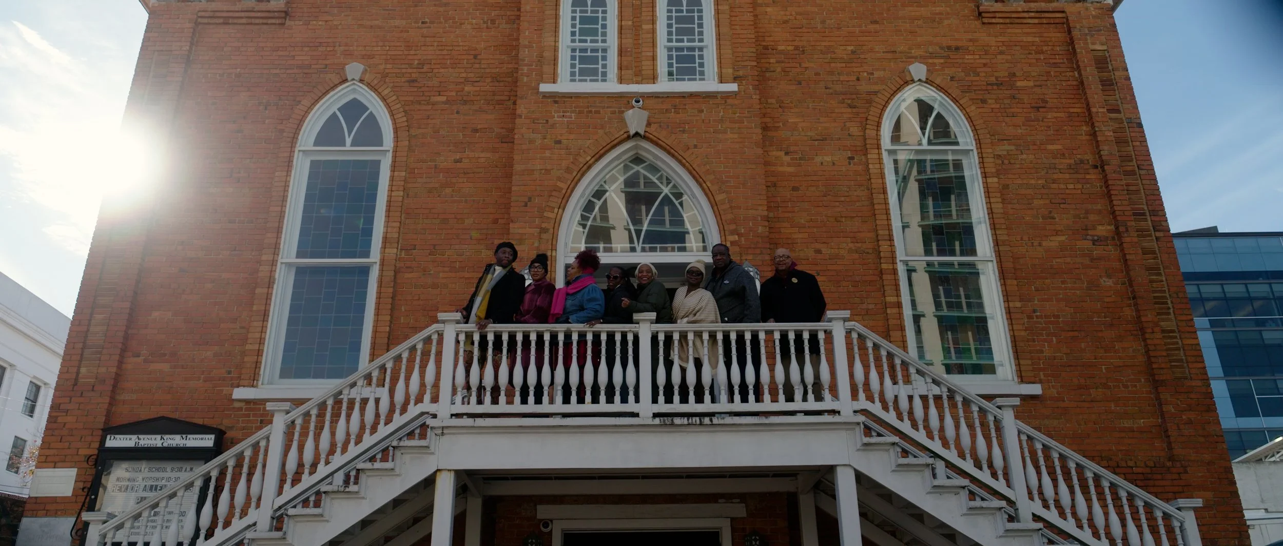 A group of people standing on a staircase in front of a brick church building with tall, arched windows.