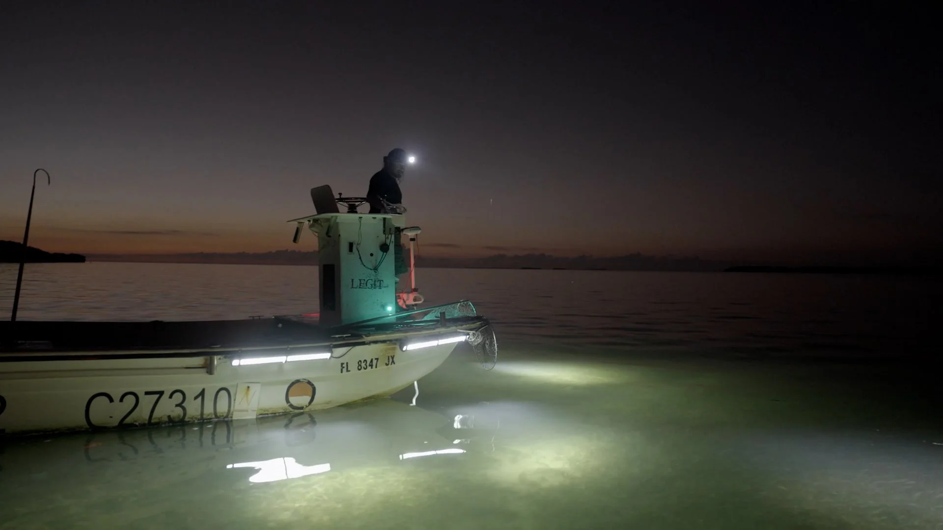 Person on a small boat at dusk with lights illuminating the water, wearing a headlamp, on a calm body of water.