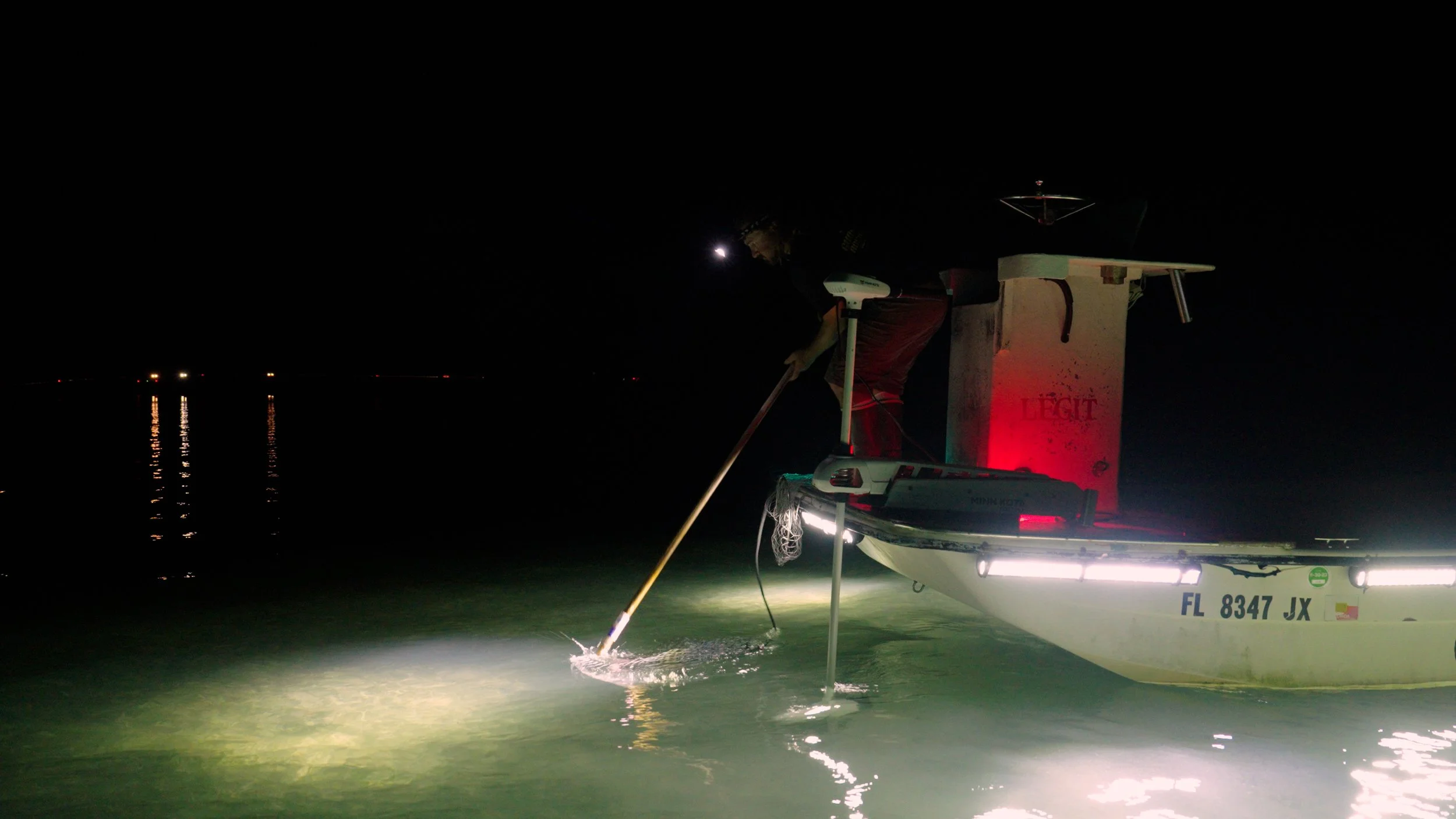 Person on a boat at night using a pole to navigate shallow water, illuminated by boat lights.