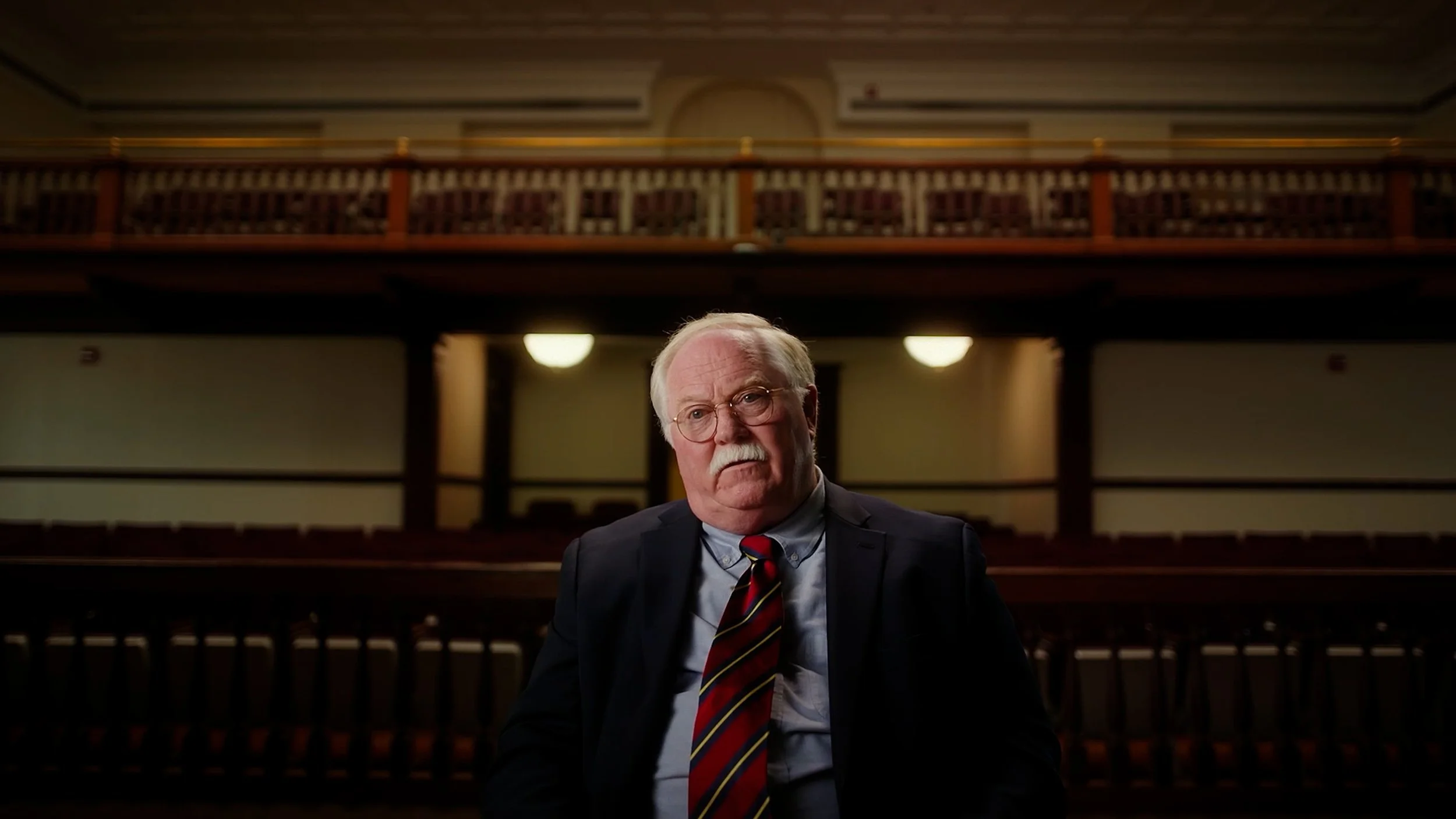 An older man with glasses and a mustache, wearing a suit and striped tie, sits inside an empty courtroom, with wooden benches and soft lighting in the background.
