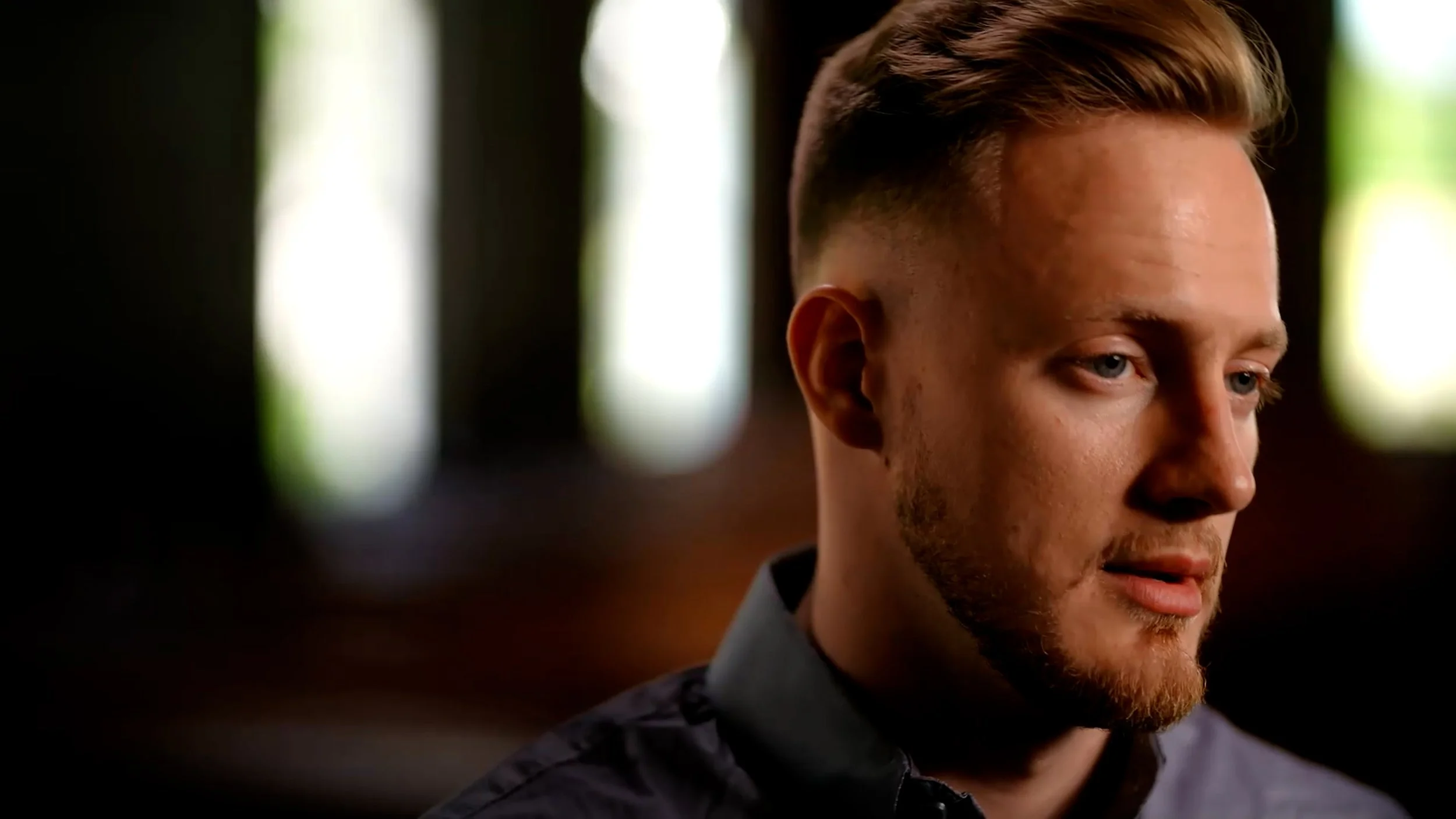 Man with short hair and beard, wearing a collared shirt, looking thoughtful, with a blurred background.