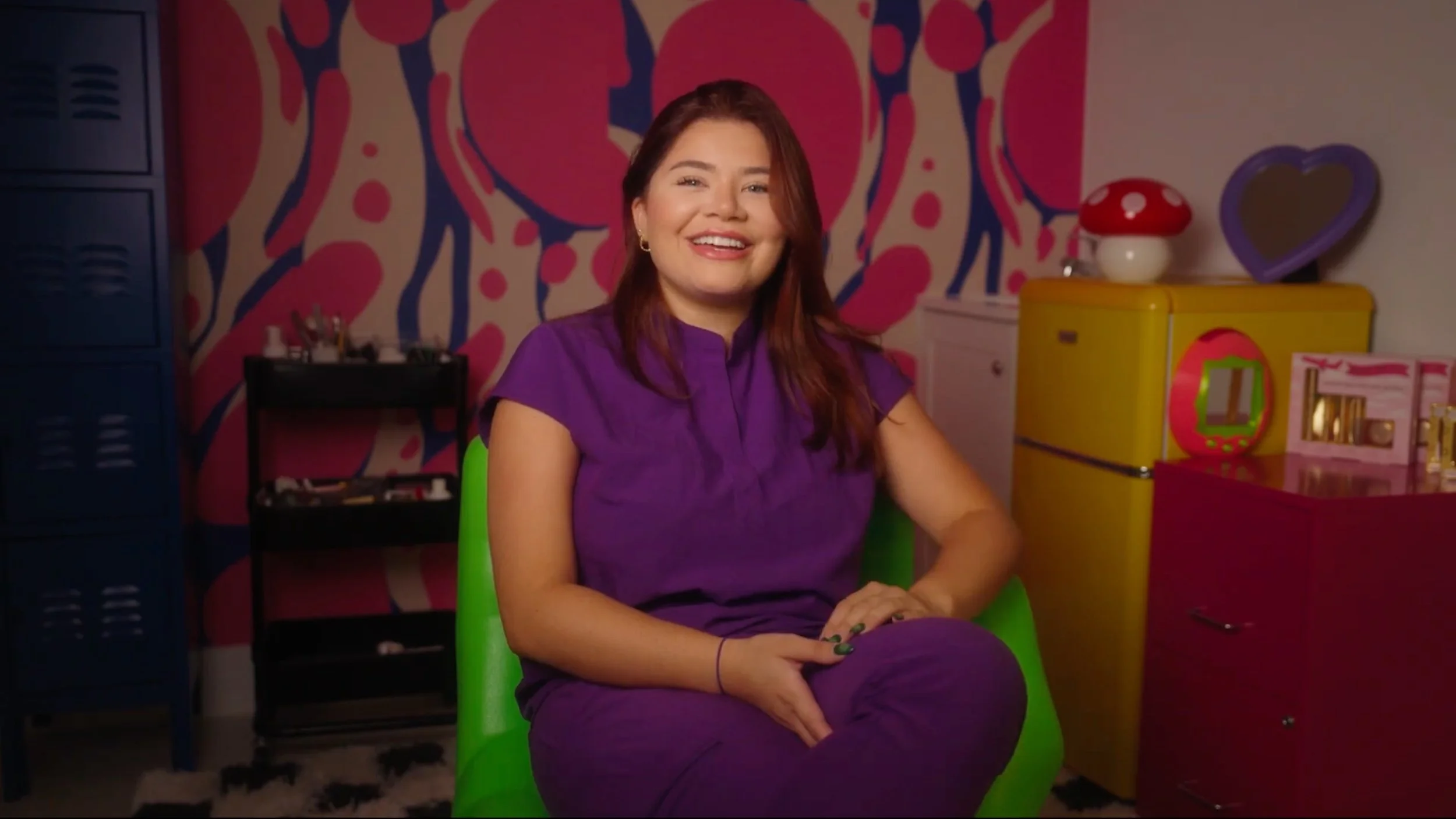 A smiling woman with reddish-brown hair wearing a purple shirt, sitting on a green chair in a colorful room with pink, yellow, purple, and blue furniture and decor.