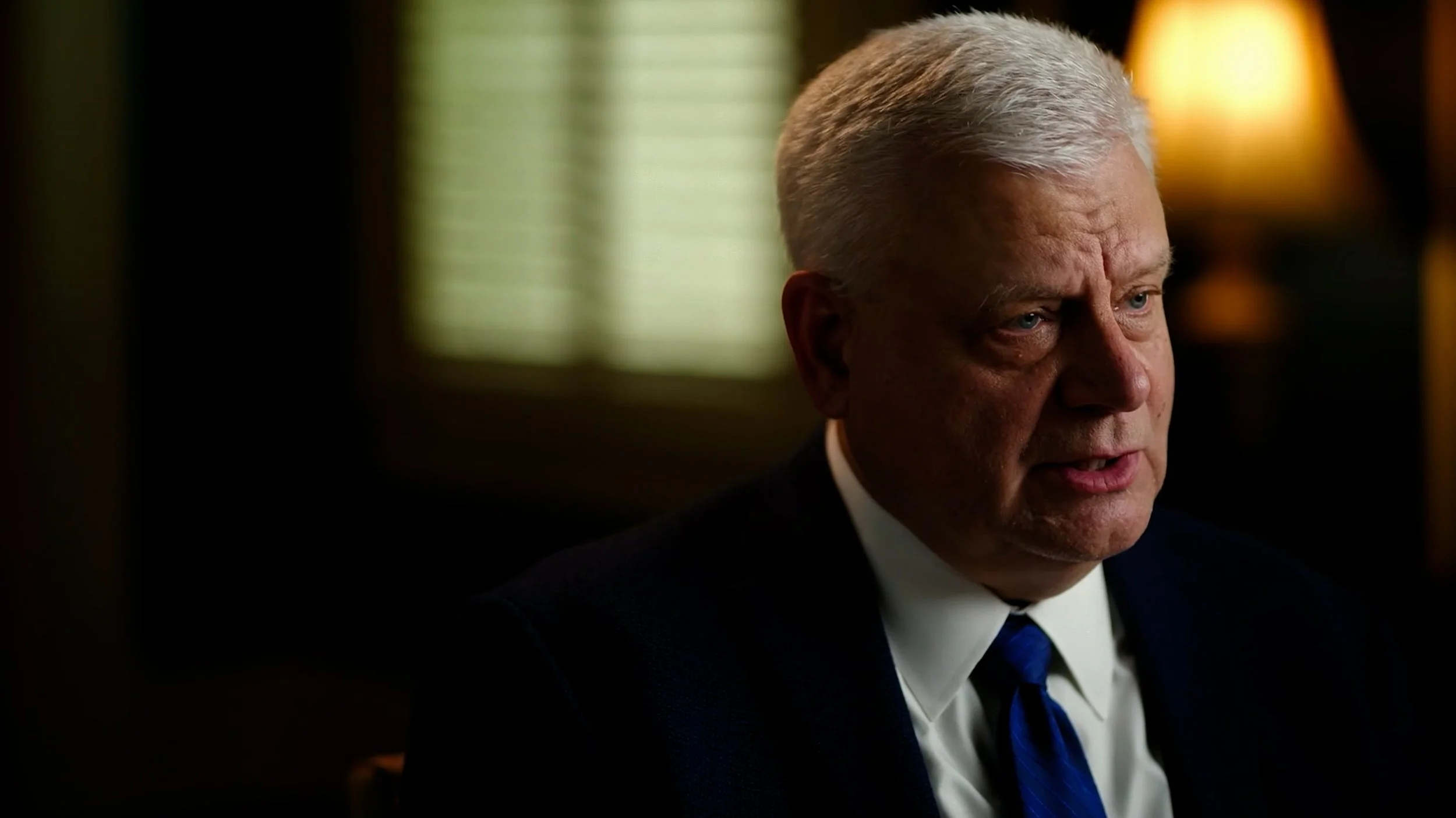 Close-up of an older man in a suit and tie speaking indoors, with a blurred background featuring a window and lamp.
