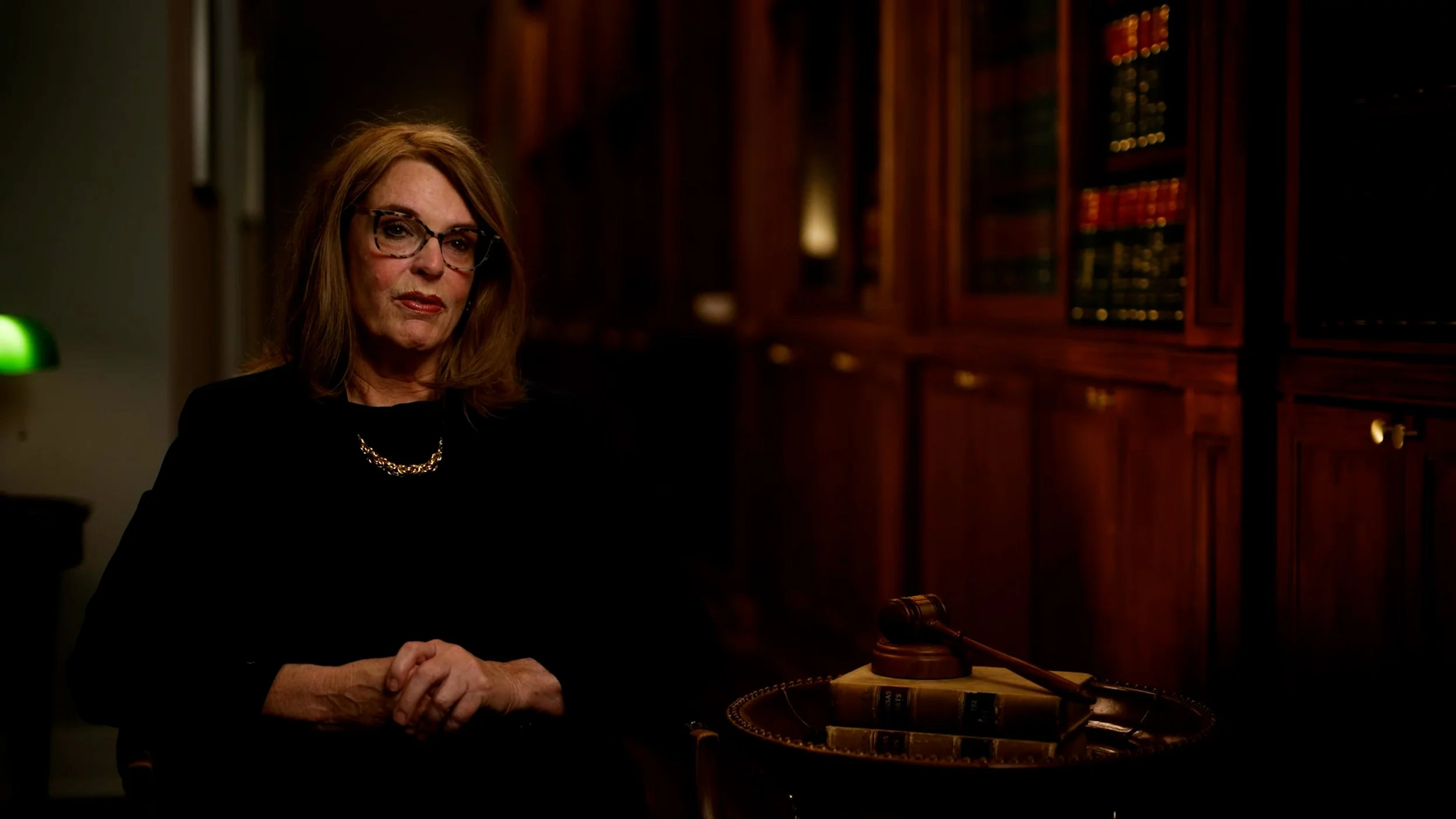 Woman sitting in a wooden-paneled room with bookshelves, wearing glasses and a black outfit, with a gavel and books on a table.