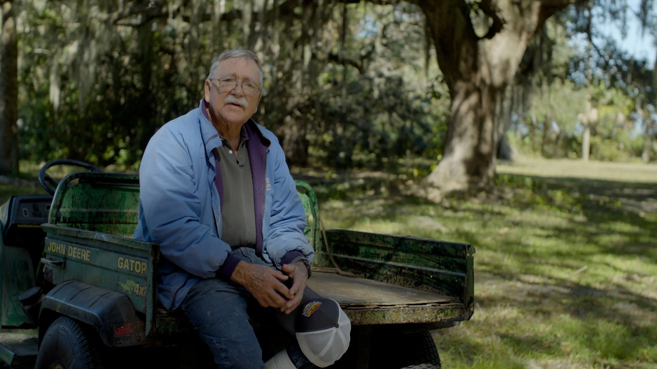 Older man sitting on a John Deere Gator utility vehicle in a forested area.