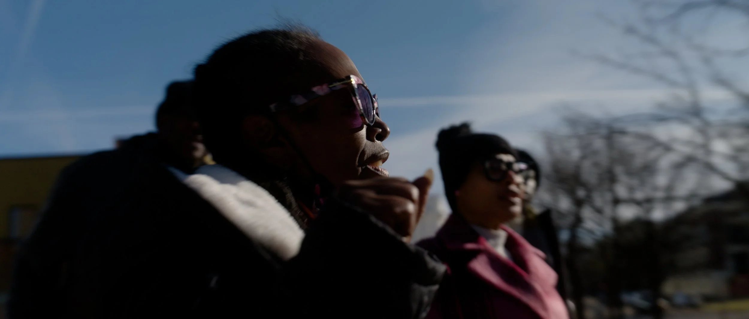 Three people standing outdoors during daytime, with a clear sky and leafless trees in the background, wearing sunglasses and warm clothing.