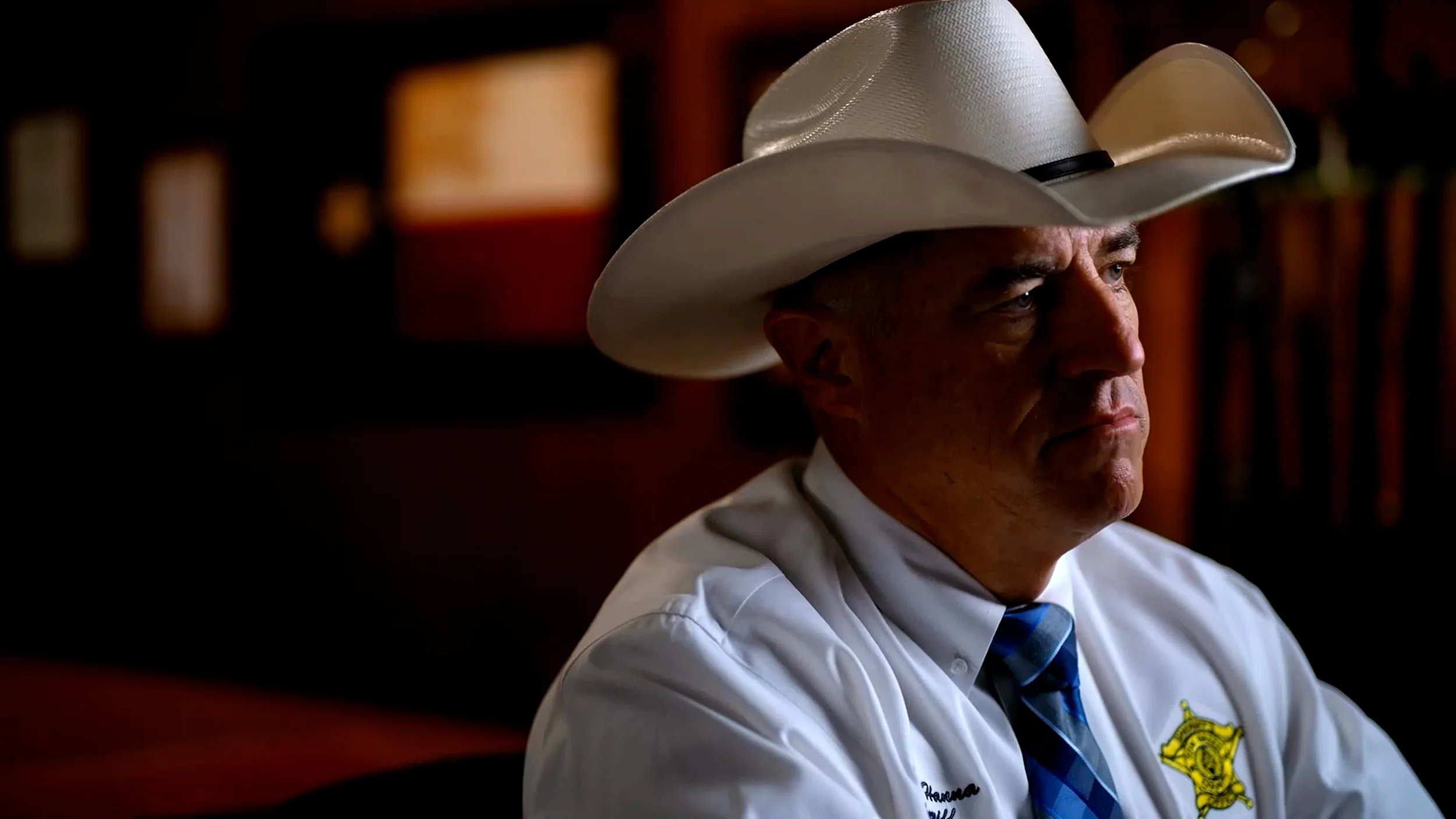 Man in a white cowboy hat wearing a white shirt with a sheriff badge emblem and a blue tie.