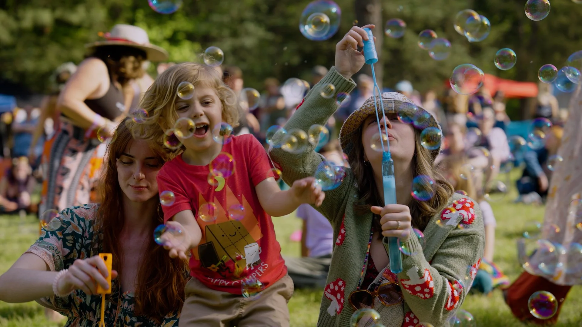 A woman and a girl blowing bubbles at an outdoor gathering with many people, trees, and a grassy area in the background.