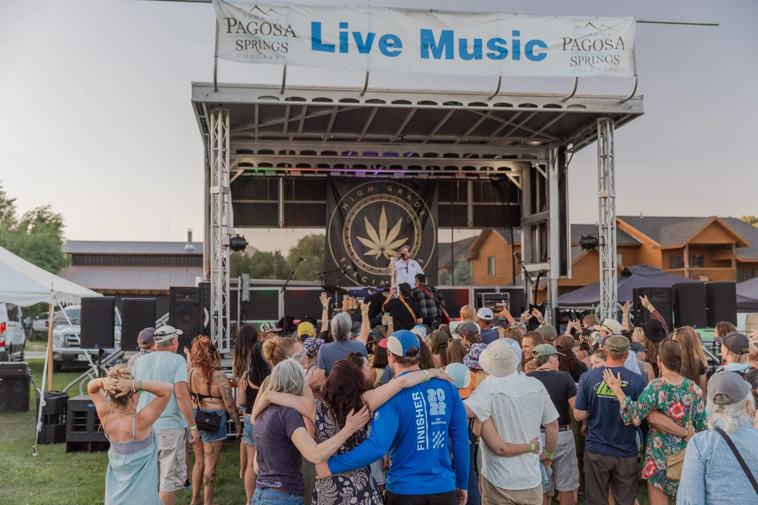 Crowd gathered close to an outdoor stage with a “Live Music” banner, watching a performer while people stand together, raise their hands, and enjoy the concert atmosphere.