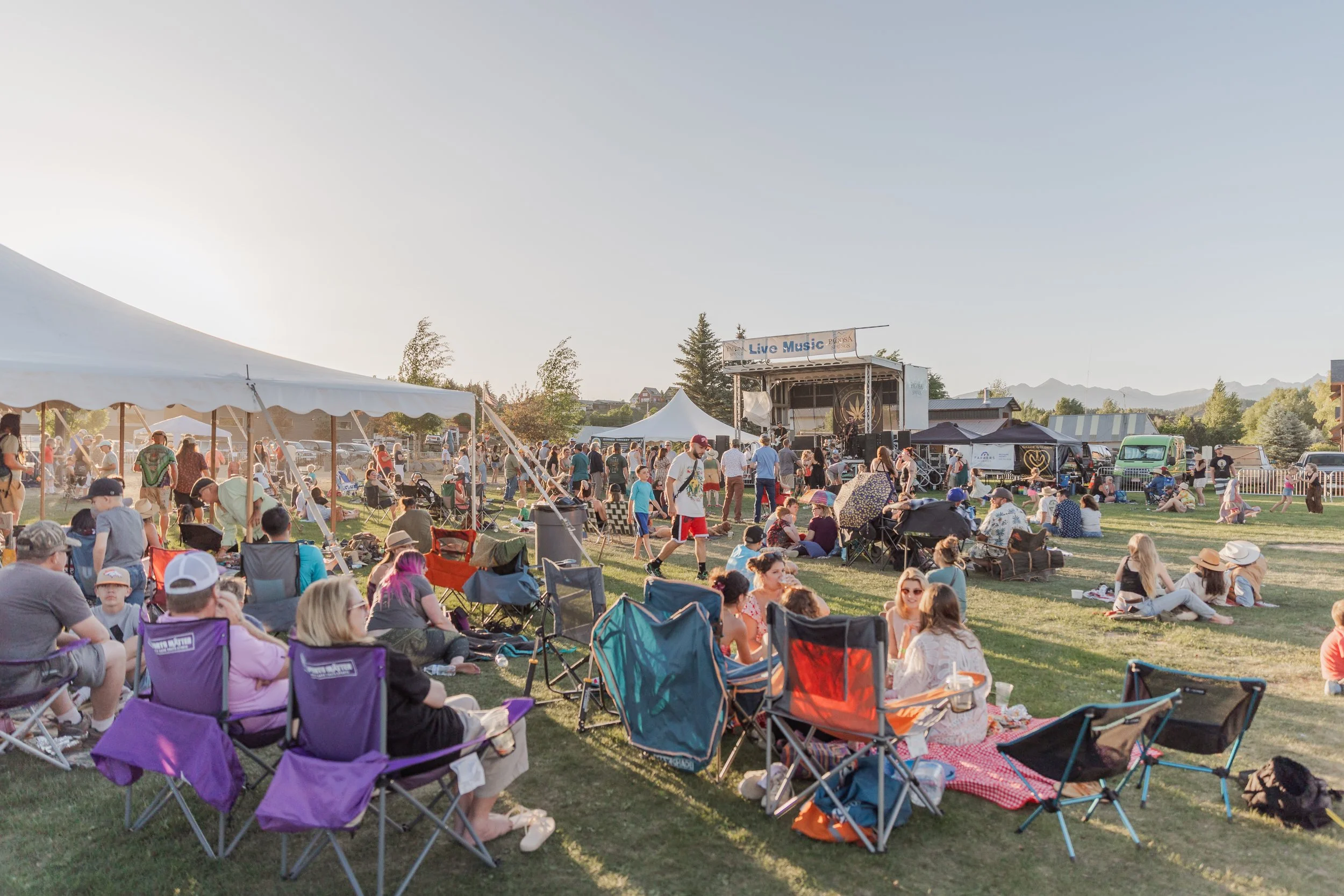 Large outdoor concert crowd gathered on a grassy field, with people sitting in lawn chairs and blankets, walking between tents, and enjoying live music on a stage in the distance during a sunny evening.