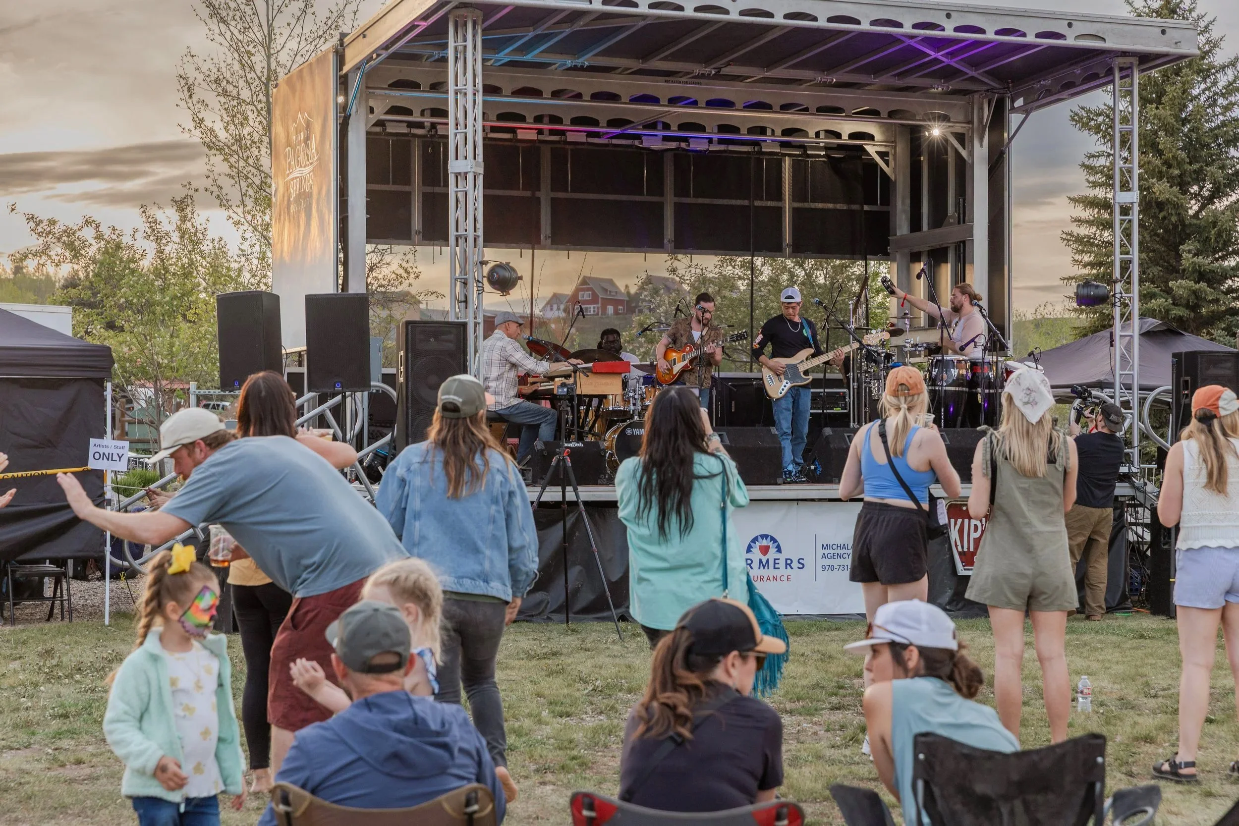 Outdoor concert at a park with a live band performing on a stage while a crowd of families and adults watches, dances, and relaxes on the grass at sunset.