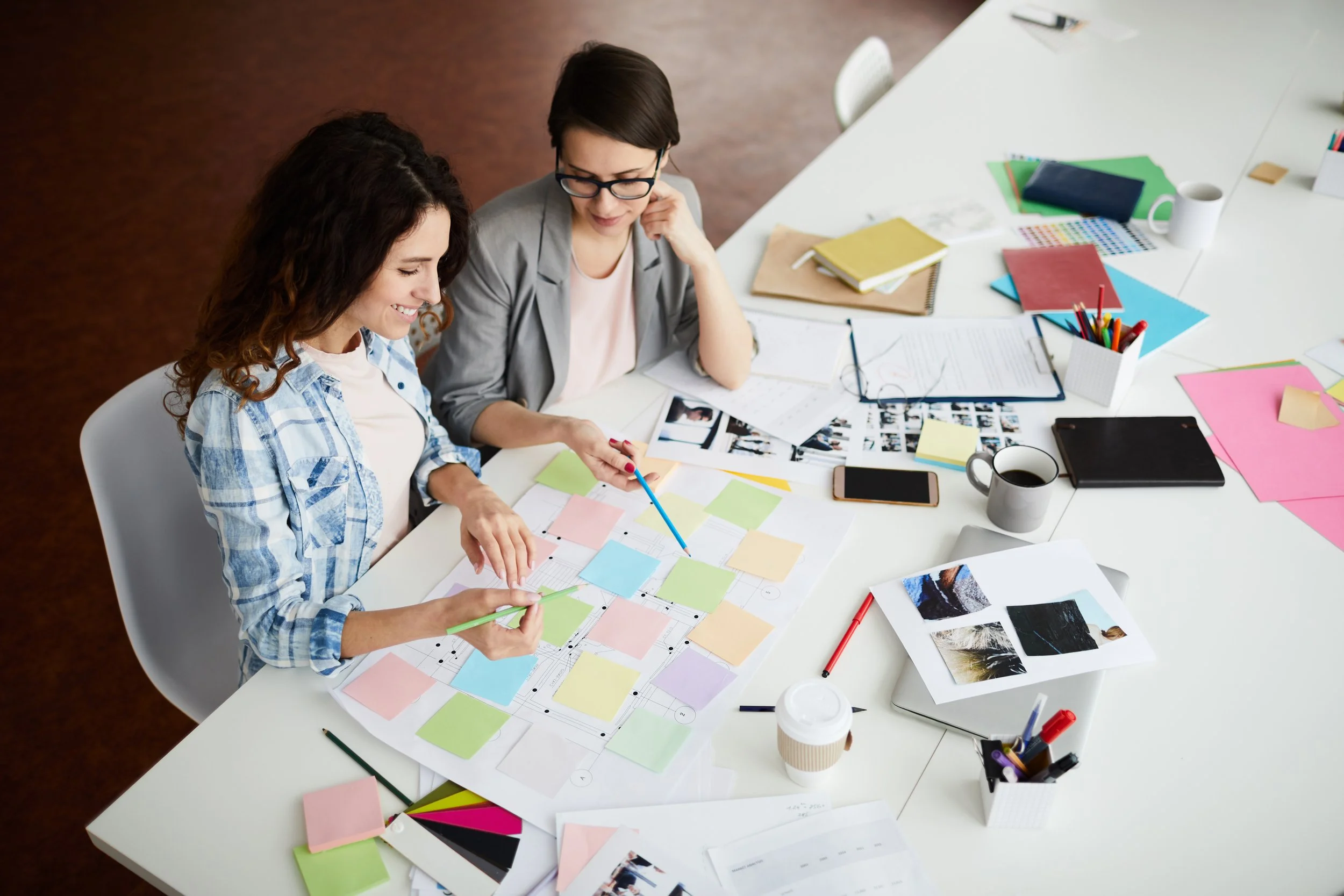 Two women working on a colorful design project at a large table, surrounded by papers, markers, and coffee cups.