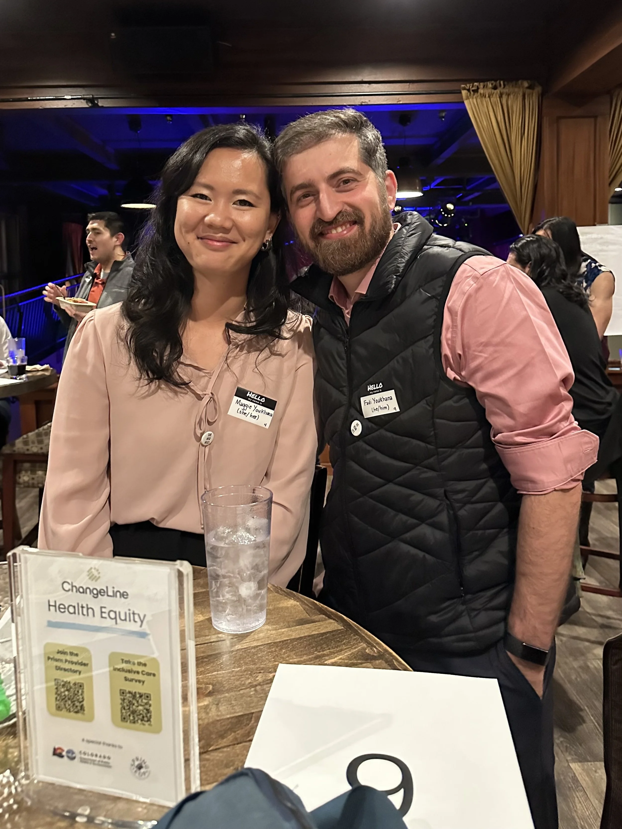  Two people standing at a table lean their heads together and smile for the camera. 