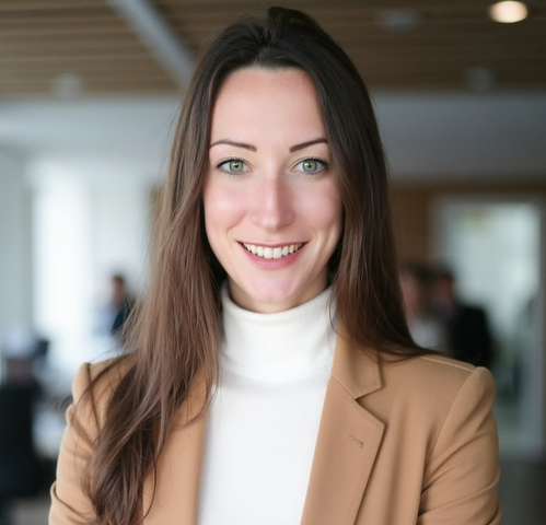 A young white woman with shoulder-length brown hair and wearing a cream turtleneck with tan blazer smiles at the camera