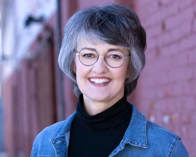 Photo of woman with short salt and pepper hair, wearing glasses and a jean jacket, smiling at the camera