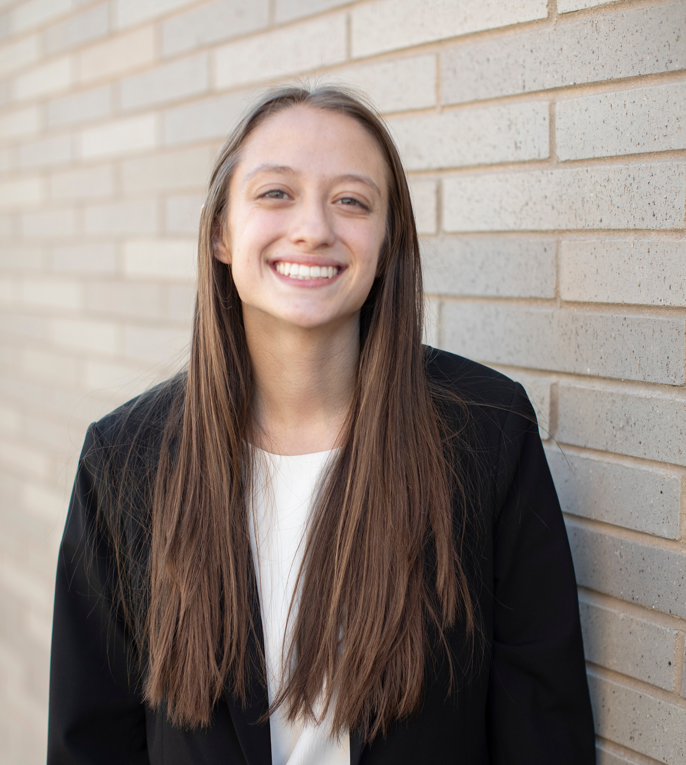 Young woman with long brown hair, in a dark blazer, smiling at camera against a gray background