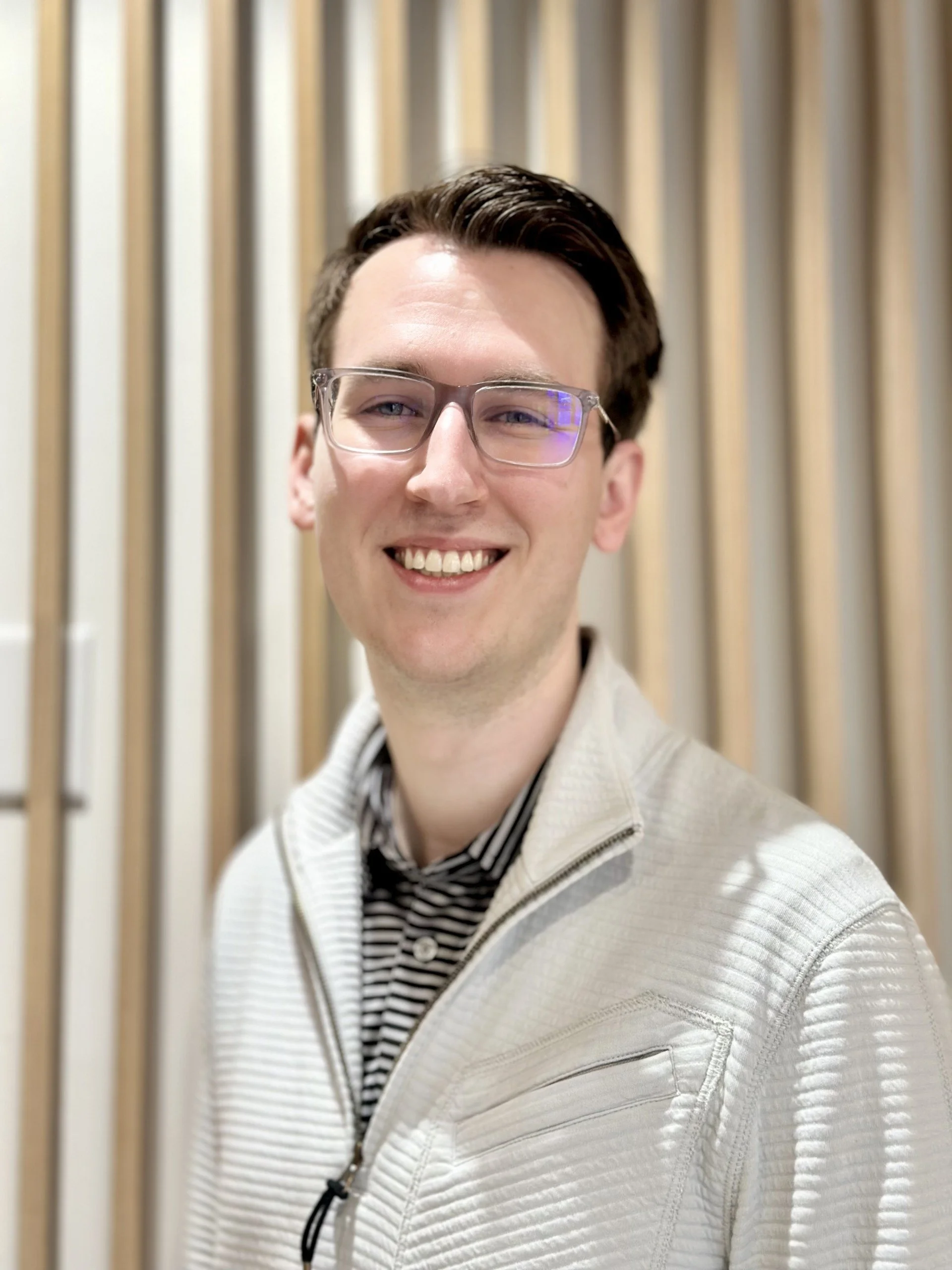 A young white man with clear-framed glasses smiles in front of a wall of light wooden slats.