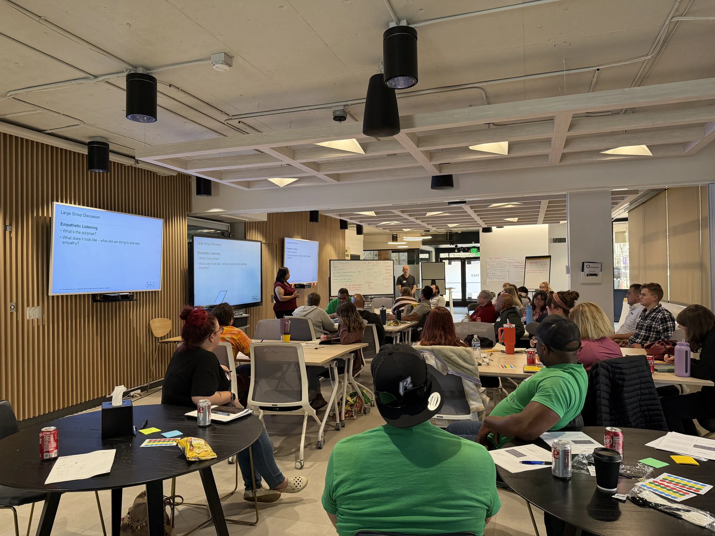 ChangeLine’s large meeting room is full of people sitting at tables watching a speaker’s presentation on three screens mounted to a wall of decorative wooden slats.