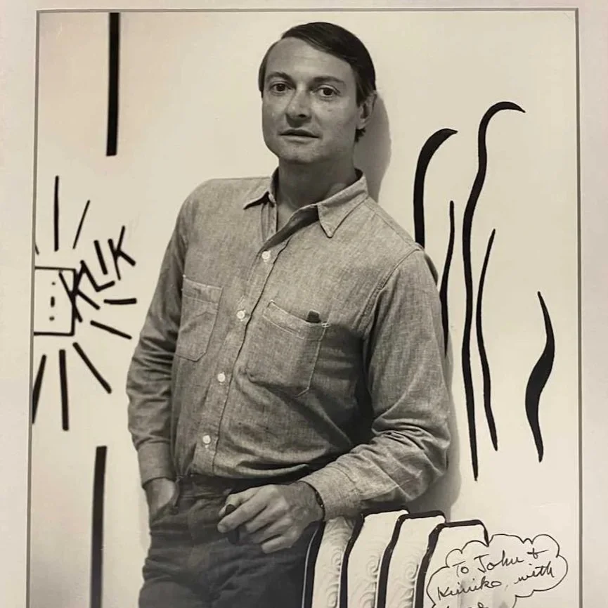A black and white photo of a man standing against a wall decorated with hand-drawn art, including the word 'CRUK' inside a box with radiating lines, and wavy lines to his right. There is also a handwritten note at the bottom right corner.