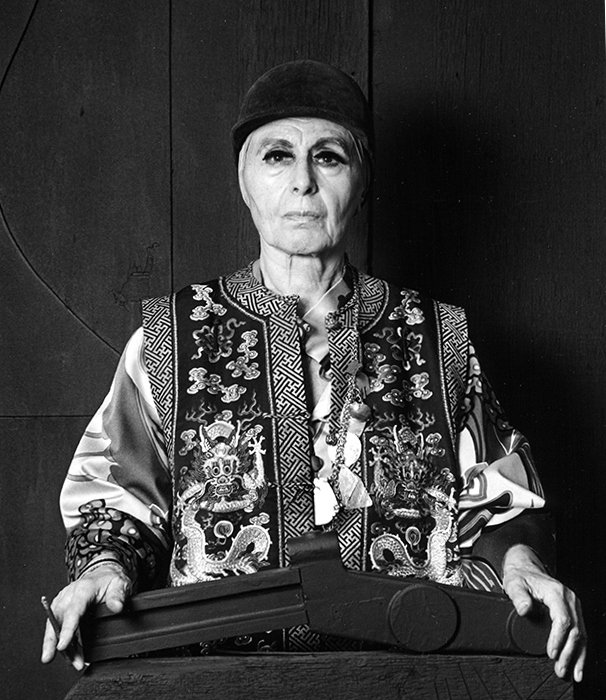 A stern-looking elderly woman in traditional Asian attire, wearing a dark head covering, standing behind a podium with a dark background.