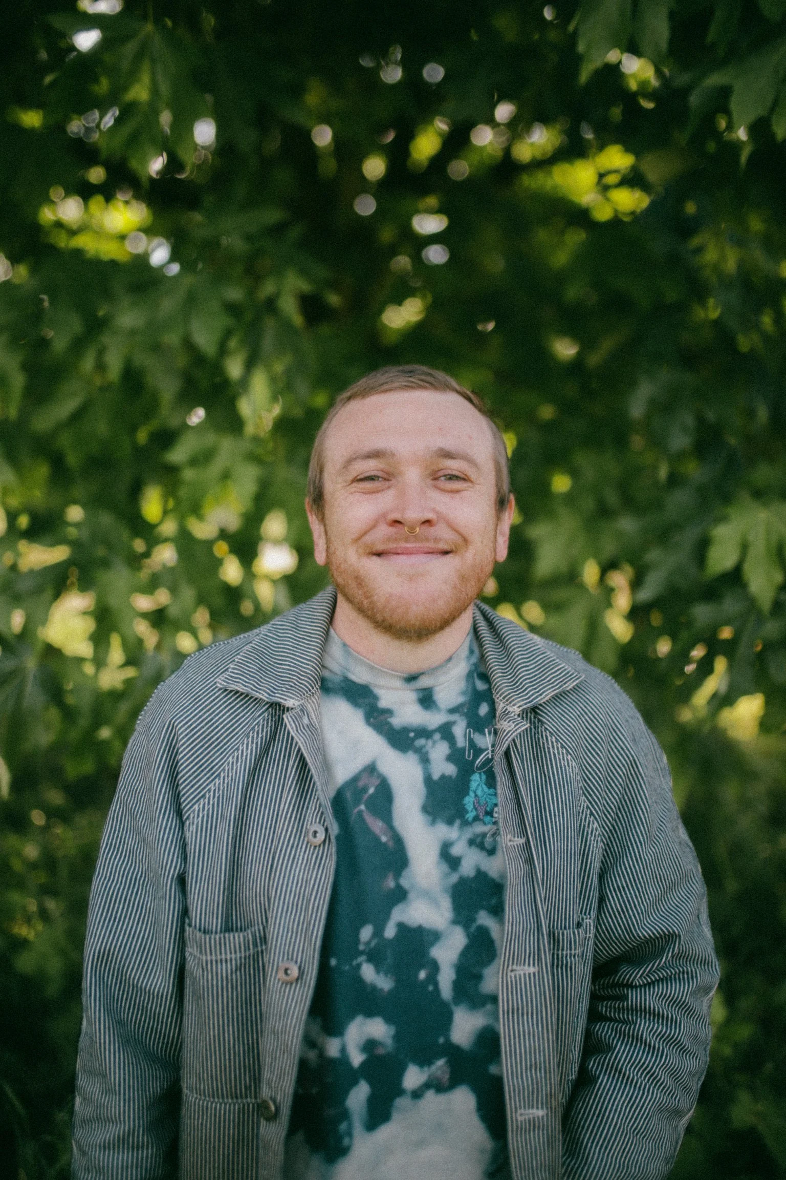 Smiling person with a septum piercing and a beard outdoors, wearing a striped jacket over a tie-dye shirt, standing in front of green leaves.