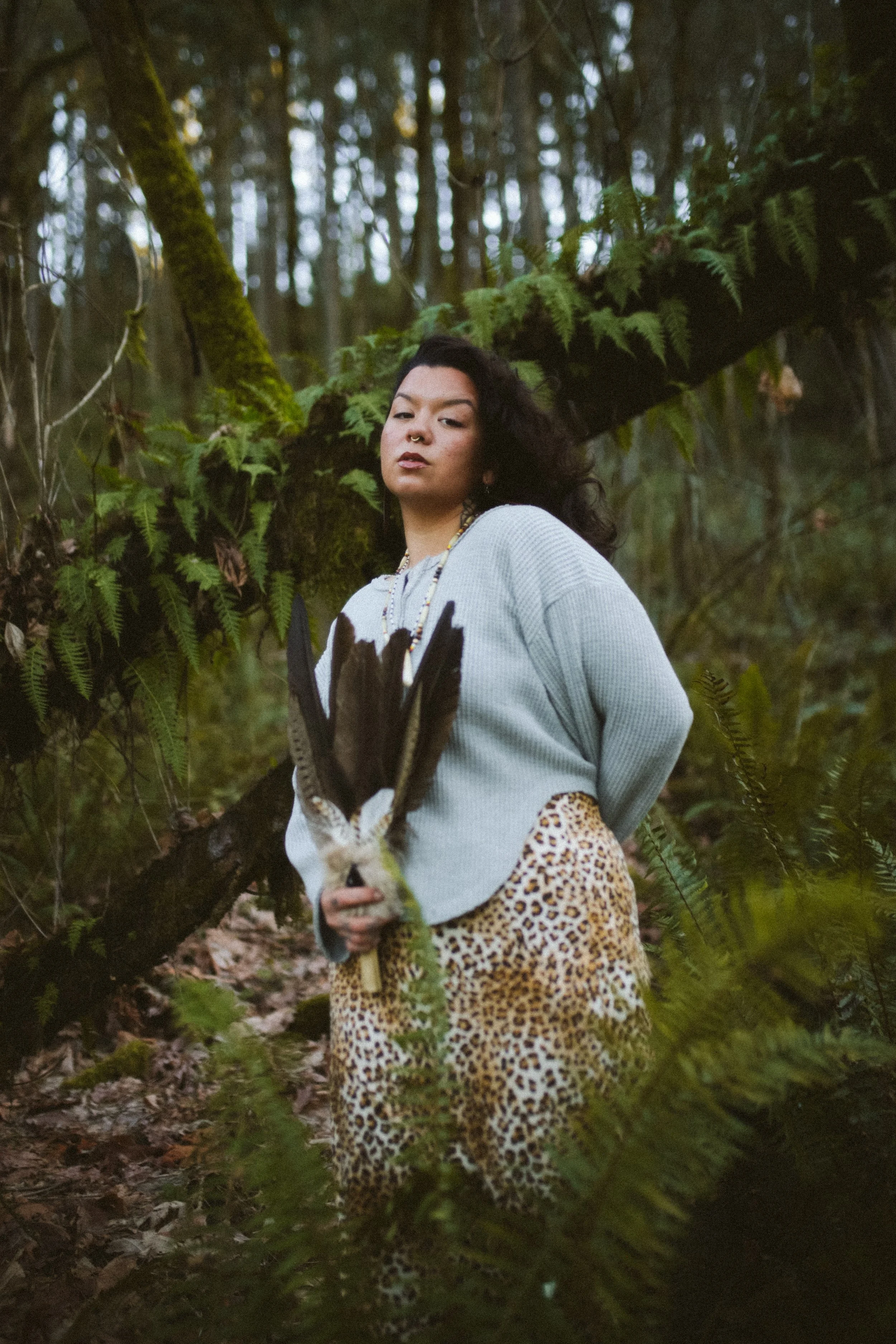 A person with dark curly hair and a septum piercing standing in a forest, holding a large feathered staff, wearing a light gray sweater and leopard print skirt.