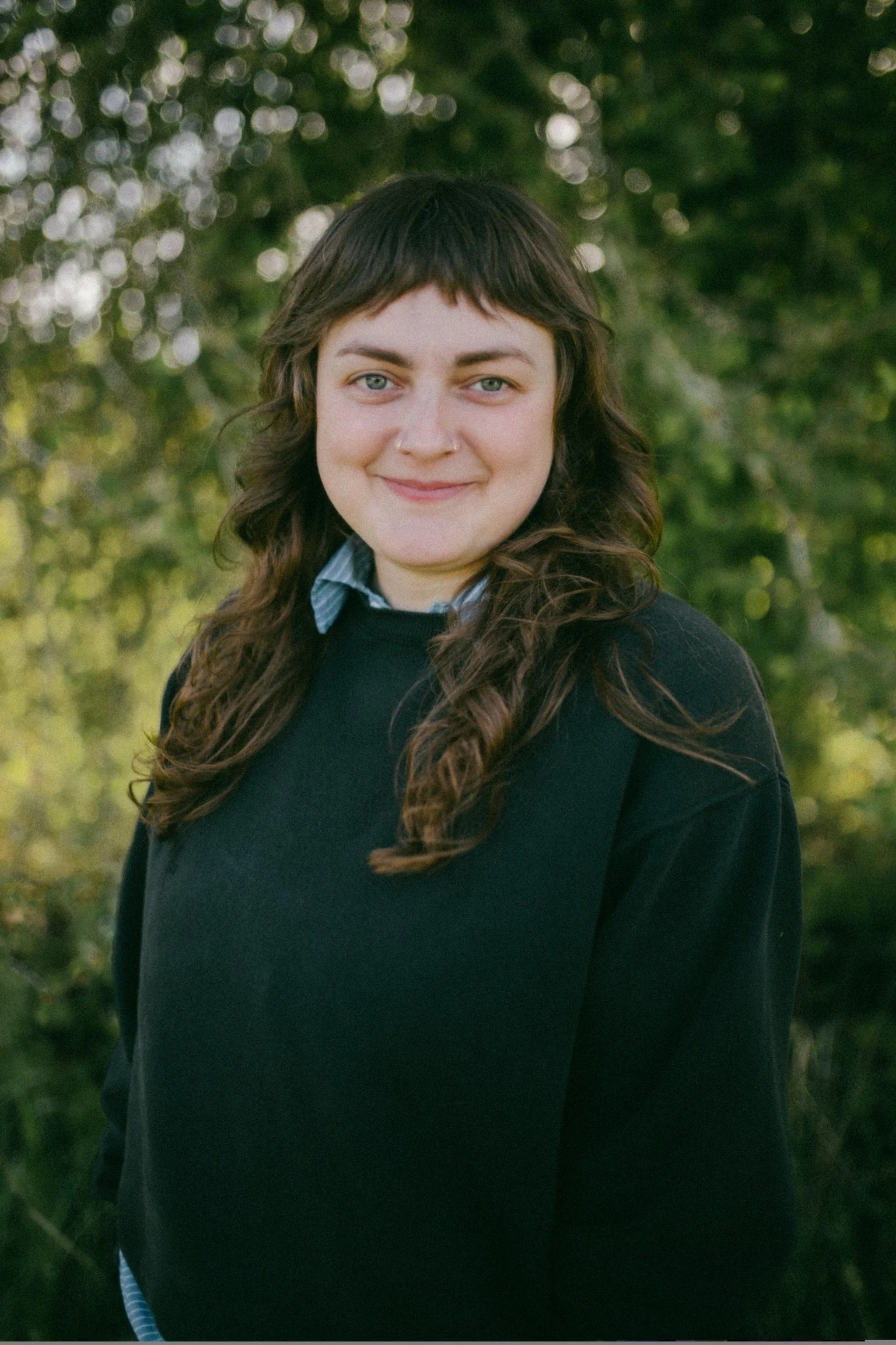 A young person with shoulder-length wavy brown hair, wearing a black sweater over a blue collared shirt, standing outdoors in front of green trees, smiling at the camera.