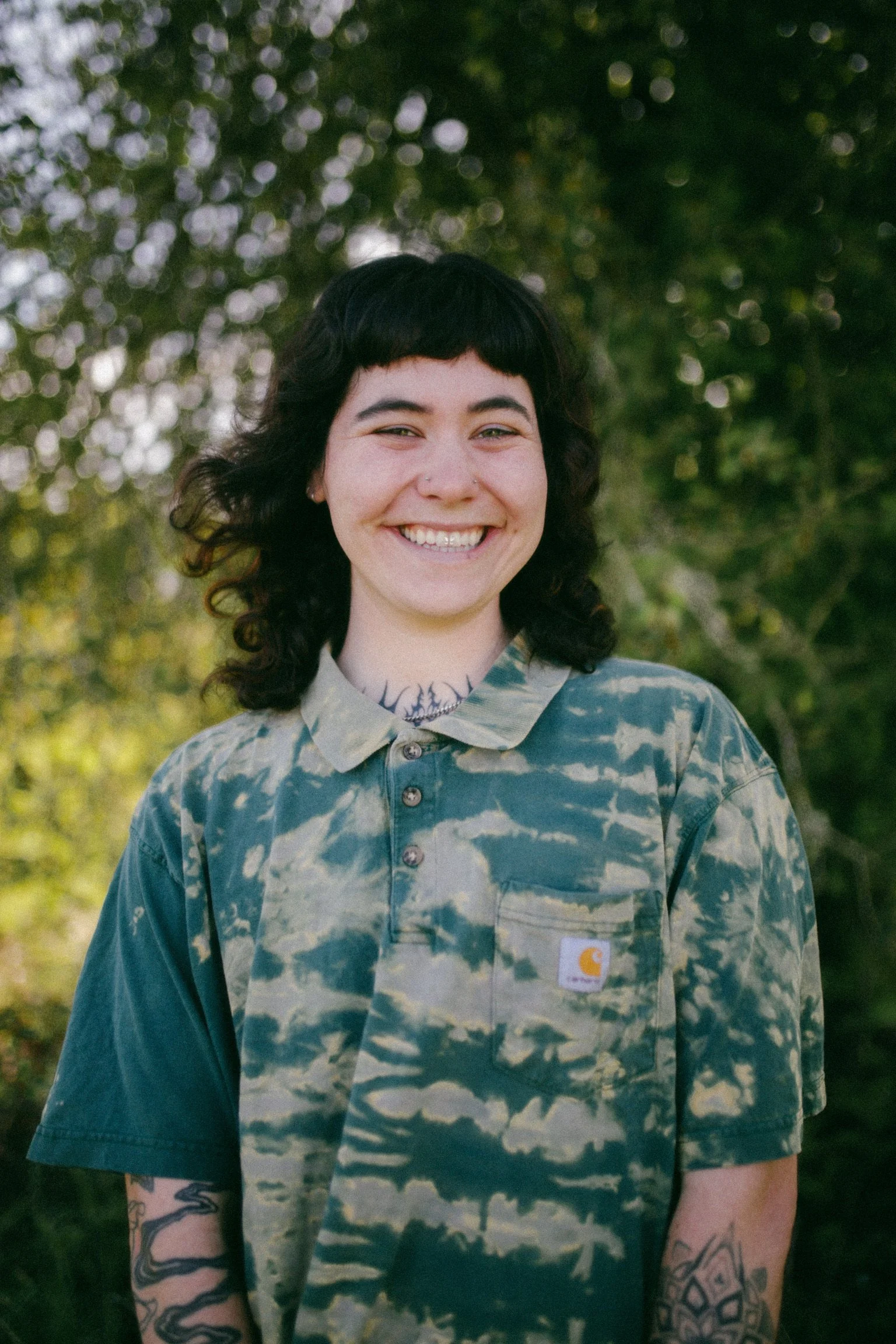 Young person with dark, curly hair and tattoos smiling outdoors during daytime, wearing a tie-dye Carhartt shirt.
